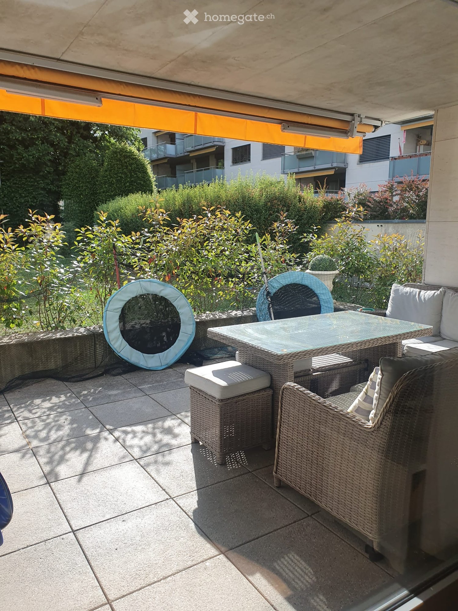Balcony with a glass table, wicker chairs, and a sofa, surrounded by greenery and a modern building in the background.