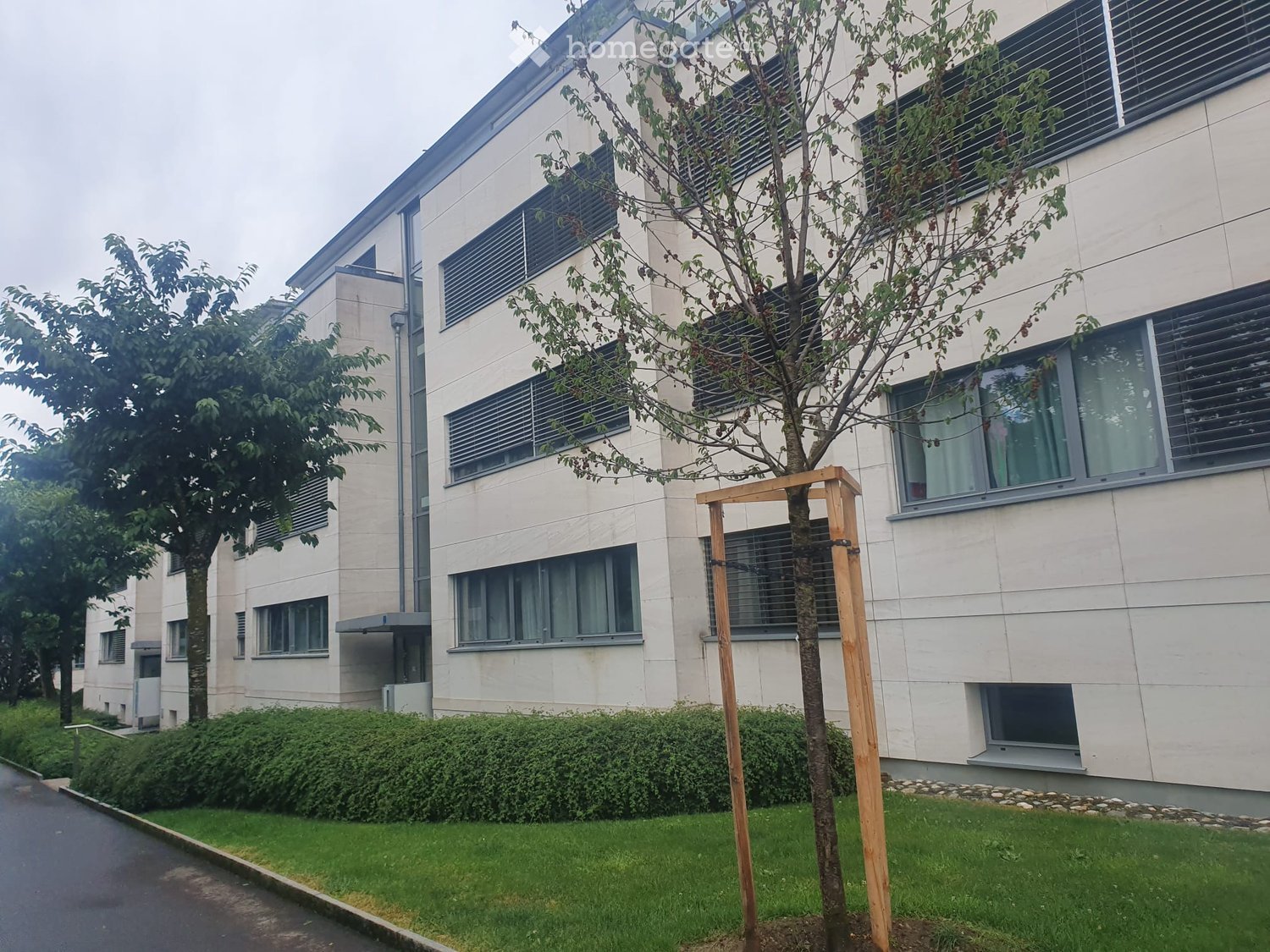 Modern multi-story building with light-colored facade, large windows, and a small landscaped area with trees and shrubs in front.