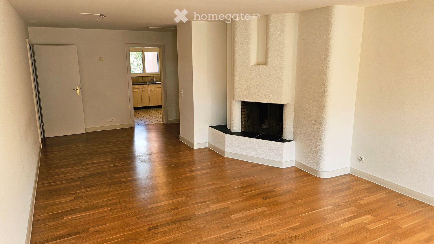 Spacious living room with polished hardwood floors, white walls, and a built-in fireplace. A doorway leads to a kitchen area with cabinets and a sink.