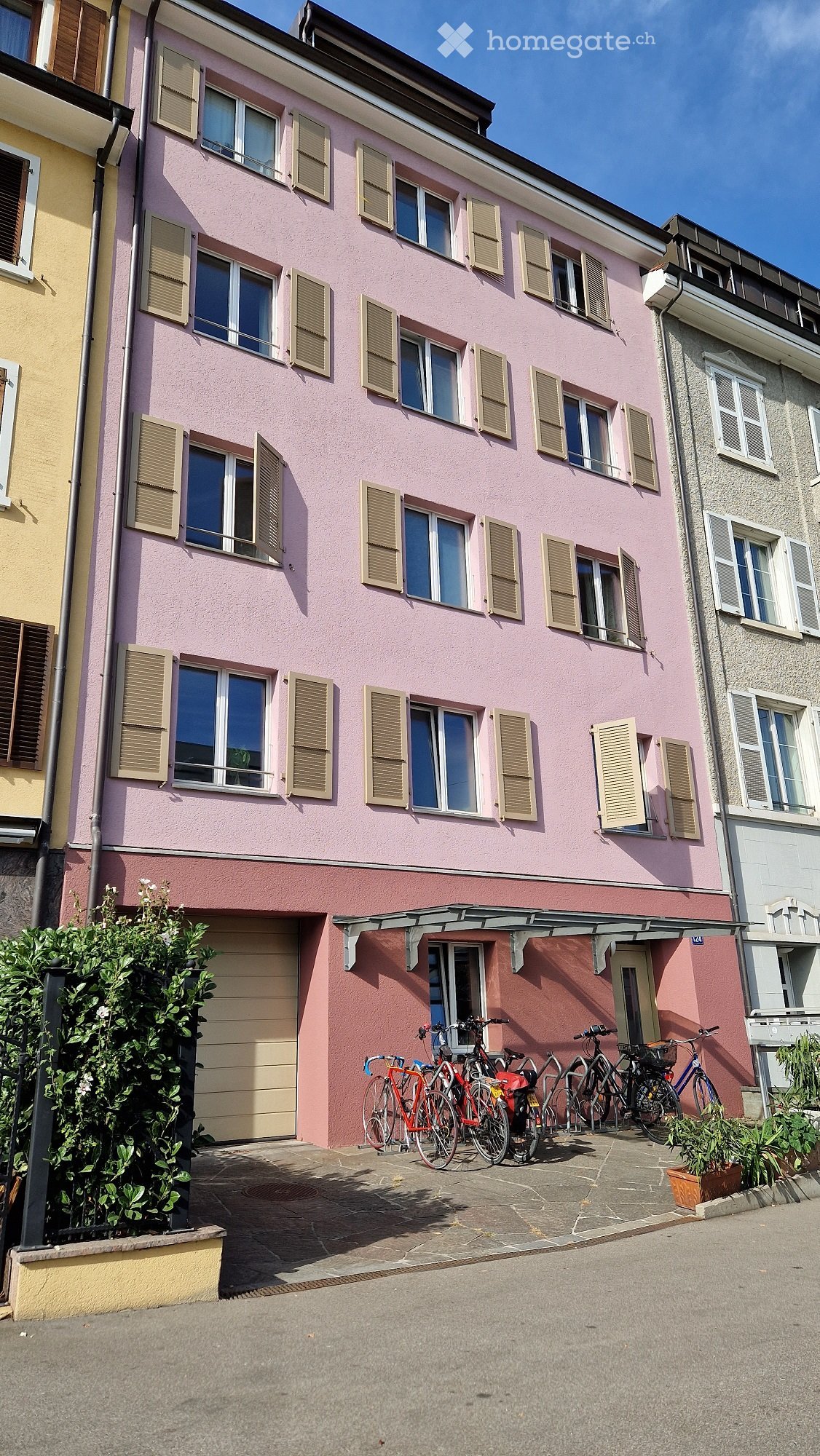 A two-story pink building with multiple windows and shutters, featuring a garage and several bicycles parked in front.