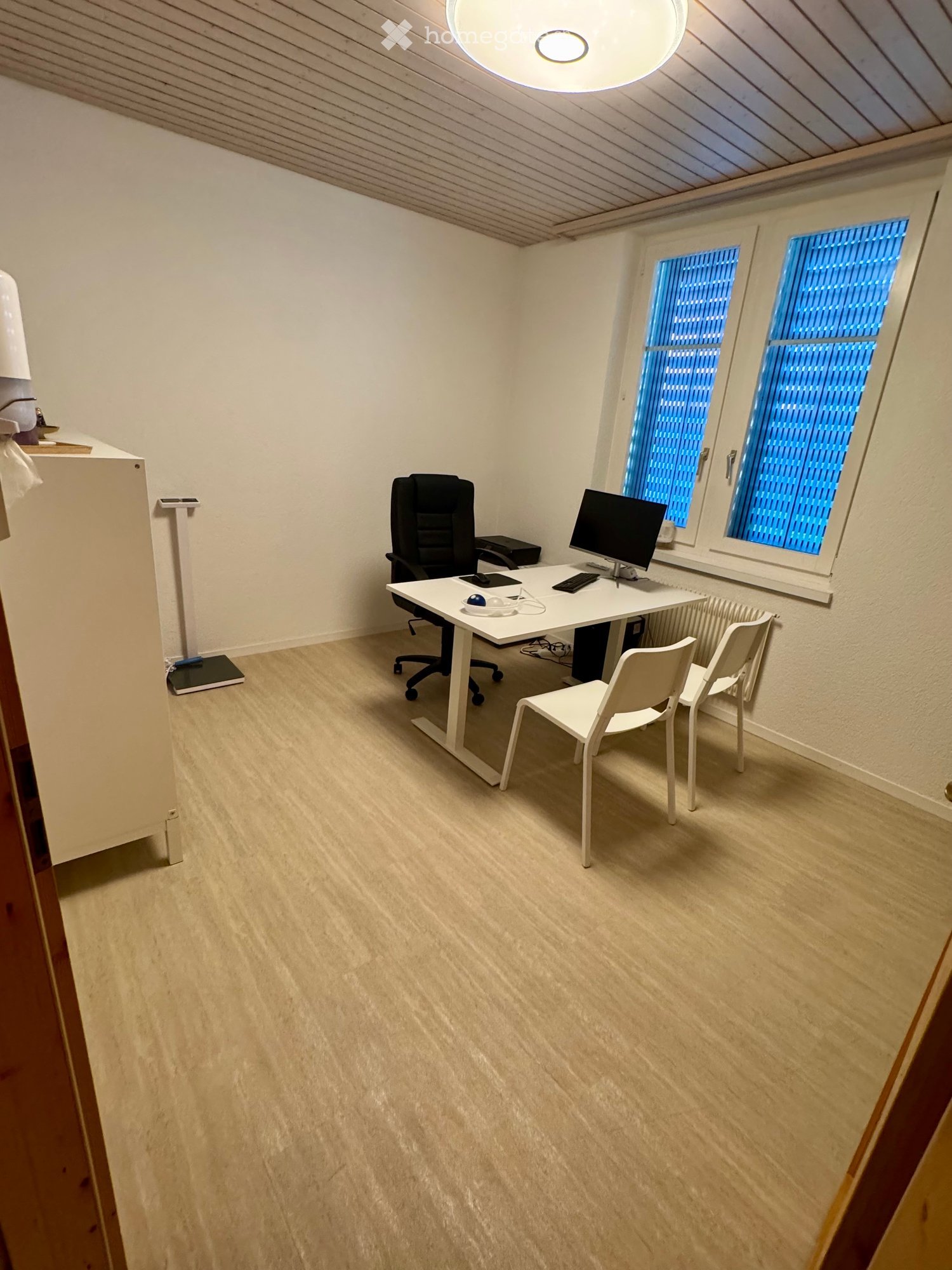 Office space with a white desk, black office chair, computer, keyboard, mouse, and two white chairs near a window with blue blinds.