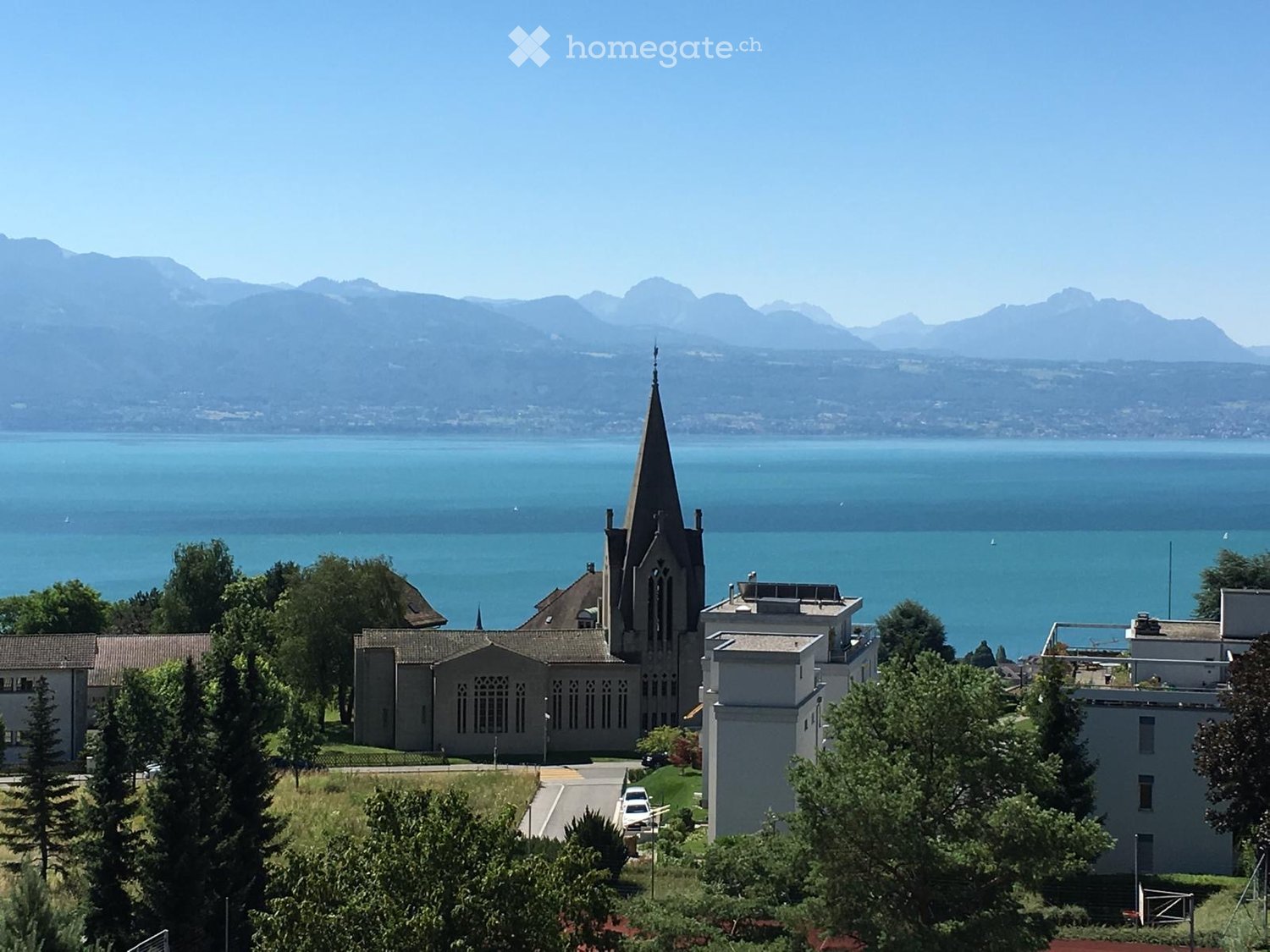 A church with a tall spire surrounded by trees, overlooking a lake and distant mountains.