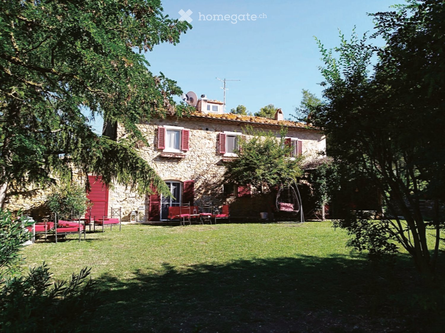A stone house with red shutters and a large, well-maintained lawn in front. Outdoor furniture including chairs and a table is arranged on the lawn, with trees providing shade.