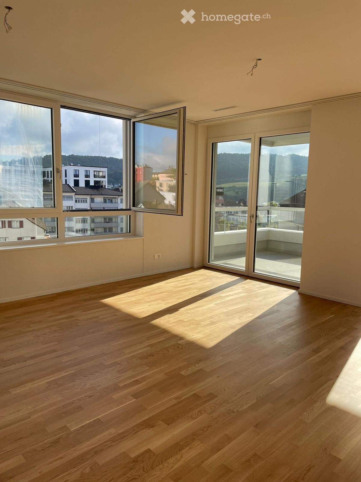 Empty room with wooden flooring, large windows, and sliding glass doors leading to a balcony.