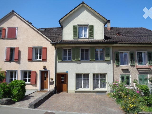 A two-story residential building with a classic European design, featuring green shutters, white walls, and a tiled roof. The entrance has a wooden door and a small garden area with plants and flowers.