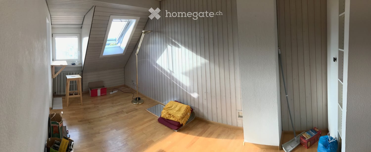 Empty attic room with wooden floors, slanted ceilings, skylight window, and minimal furnishings including a stool, floor lamp, and some boxes.