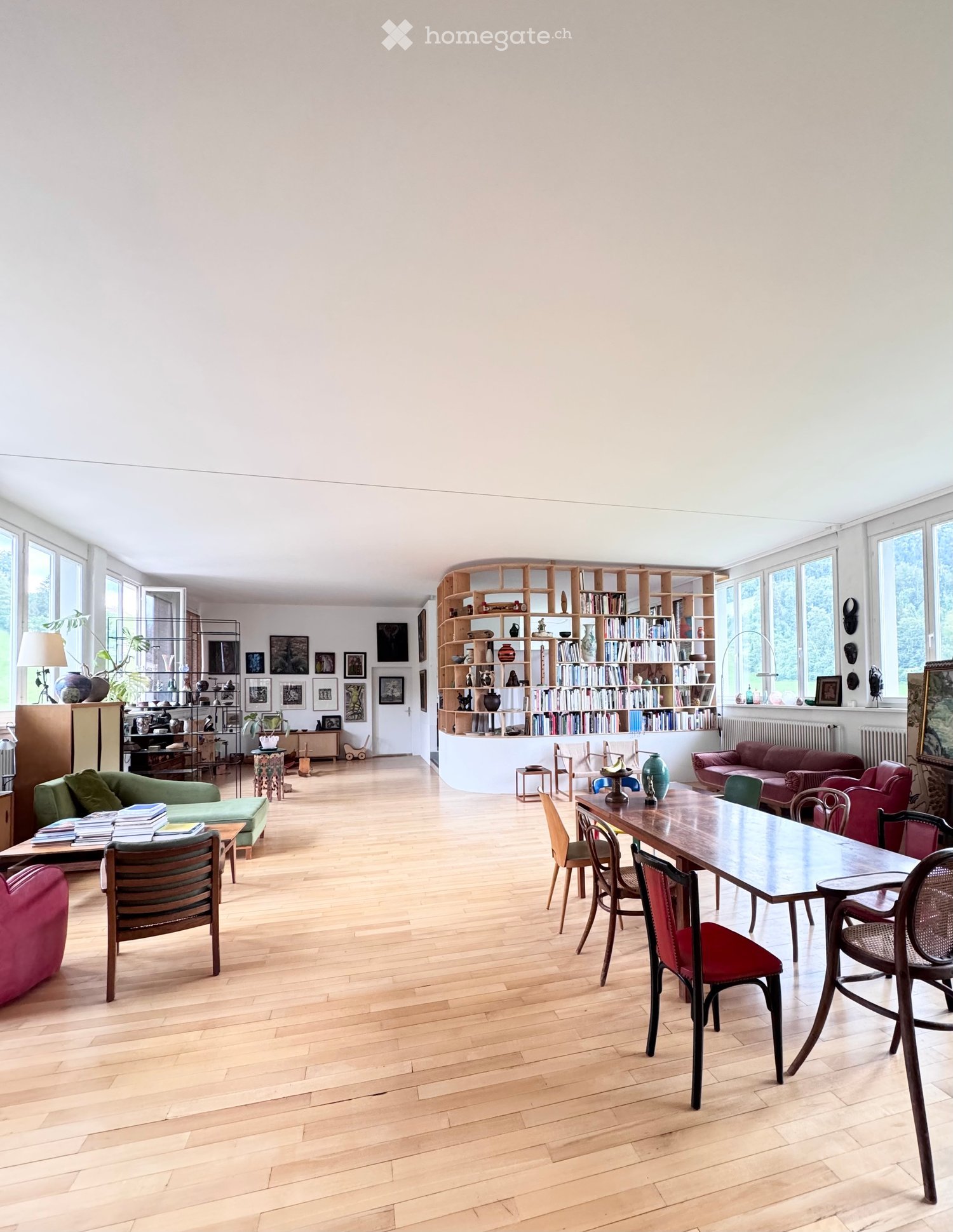 Bright living room with wooden flooring, large windows, a red sofa, a wooden dining table with chairs, a bookshelf, and decorative items.