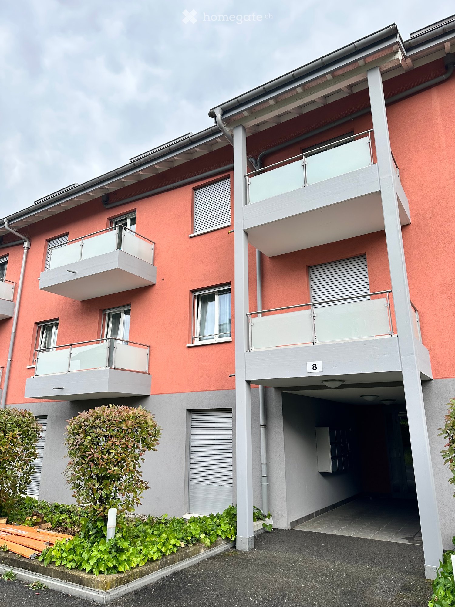 Modern two-story apartment building with pink facade, white balconies, and a covered parking area.