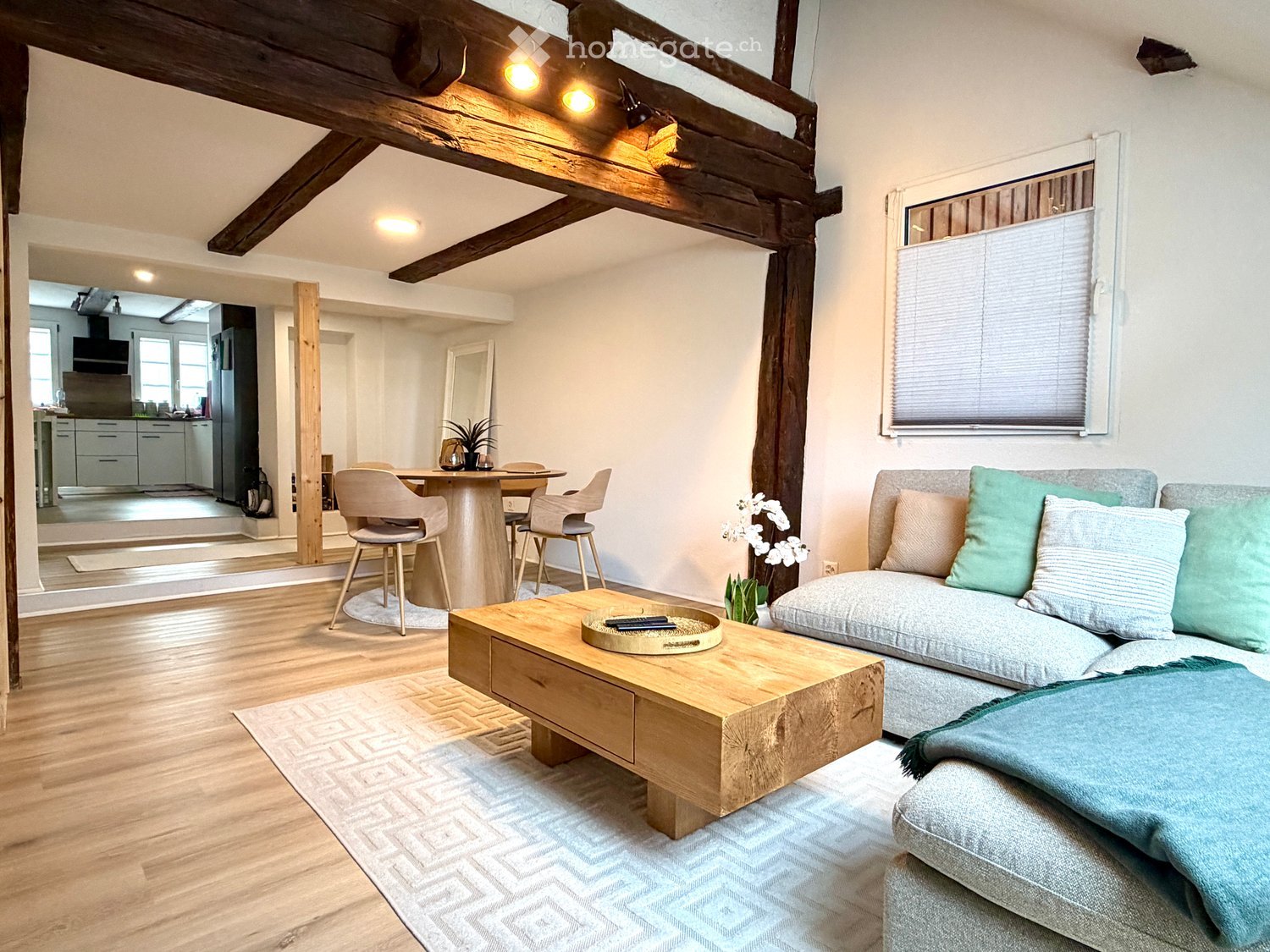 Modern living room with exposed wooden beams, a grey sofa with green and white cushions, a wooden coffee table, a dining area with a round table and chairs, and a view into a kitchen area.