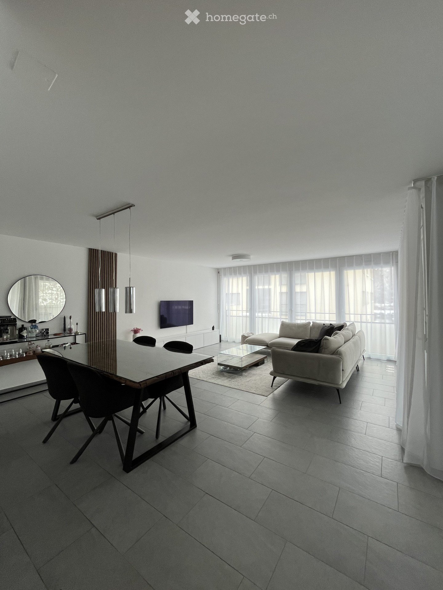 Furnished Room. Modern living and dining area with a grey sofa, coffee table, dining table with chairs, a wall-mounted TV, and a sleek mirror on a sideboard.