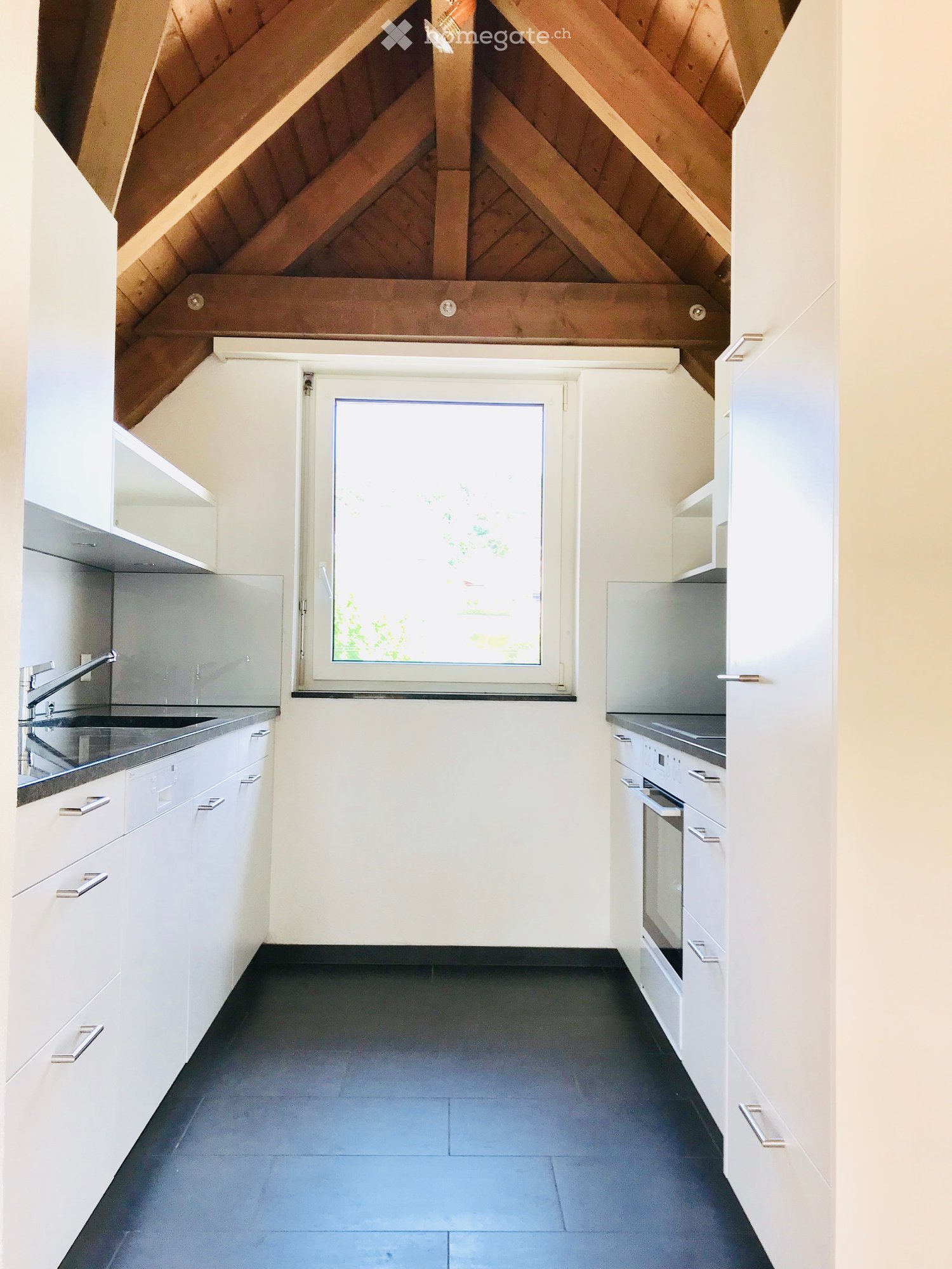 Modern kitchen with white cabinets, stainless steel sink, and oven. Features a large window and dark tiled floor.