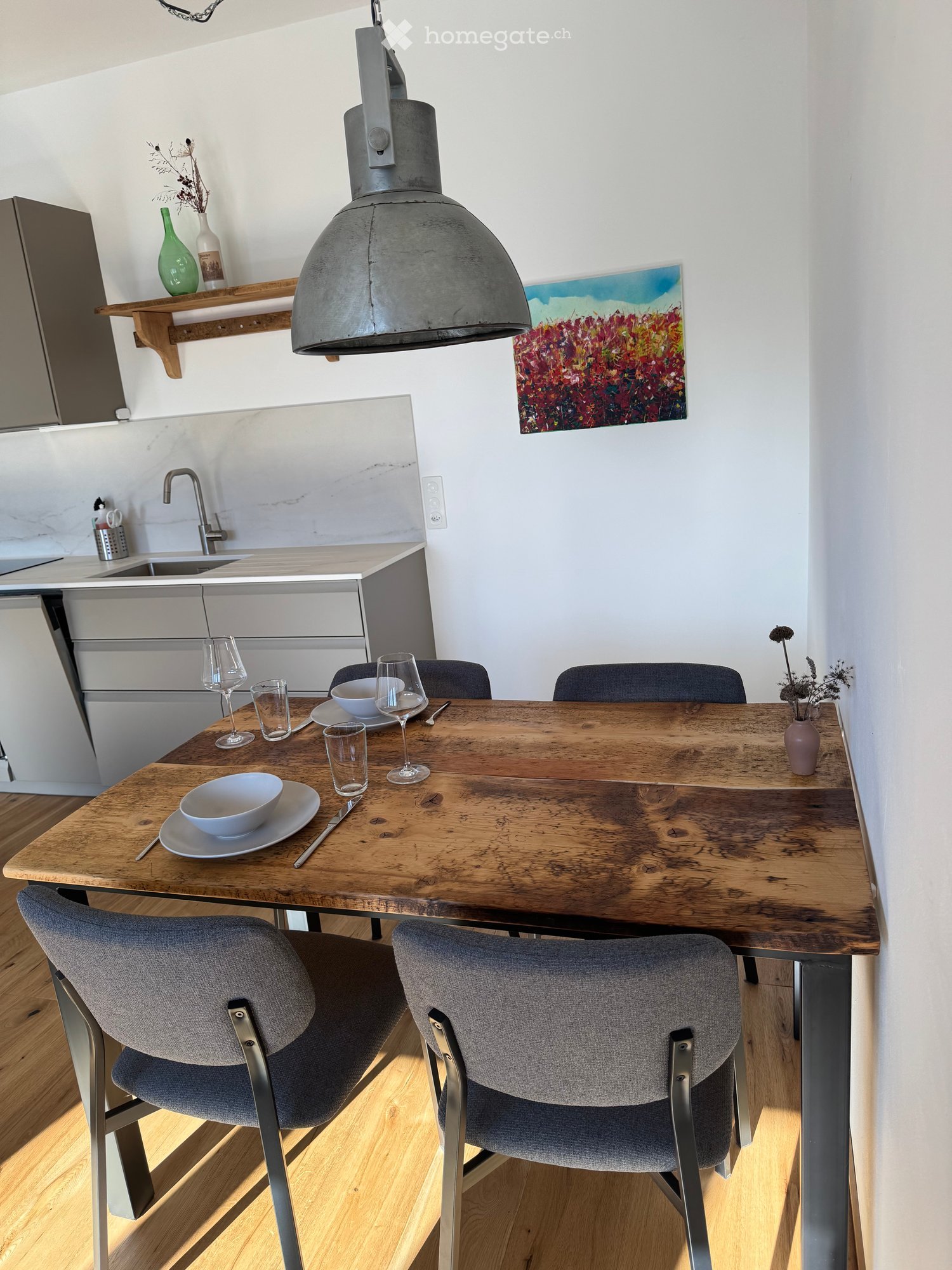 Modern kitchen with a wooden dining table, four chairs, a sink, and a pendant light. The table is set with plates, bowls, and wine glasses. A vase with dried flowers and a painting are on display.