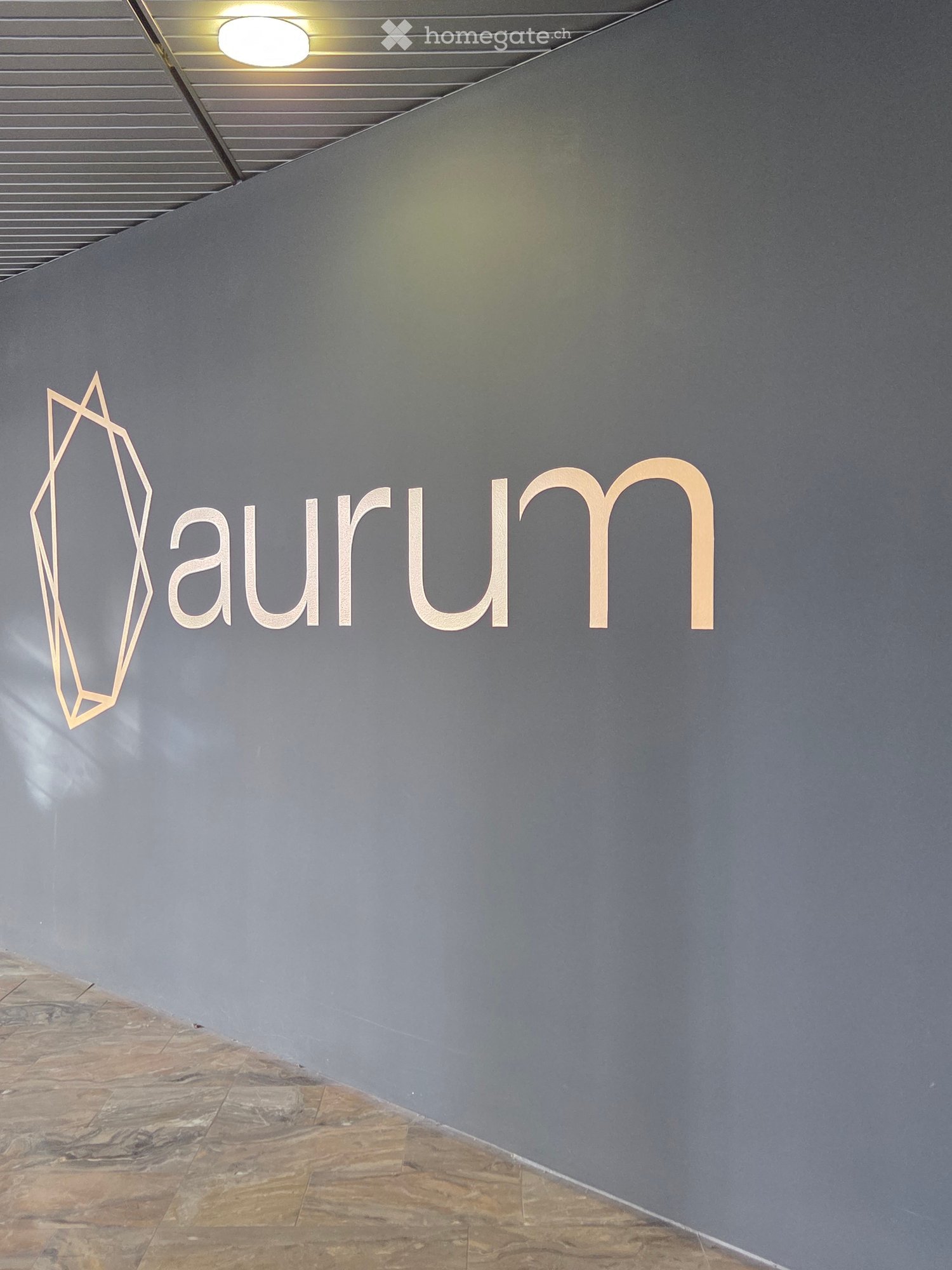 Modern hallway with grey walls, polished stone floor, and illuminated ceiling light. A geometric logo and the word "aurum" are displayed on the wall.