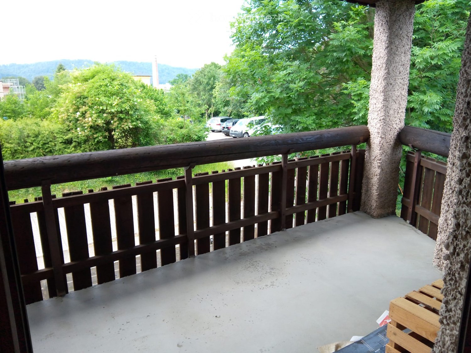 Balcony with wooden railing, concrete floor, and scenic view of trees and parked cars in the background.