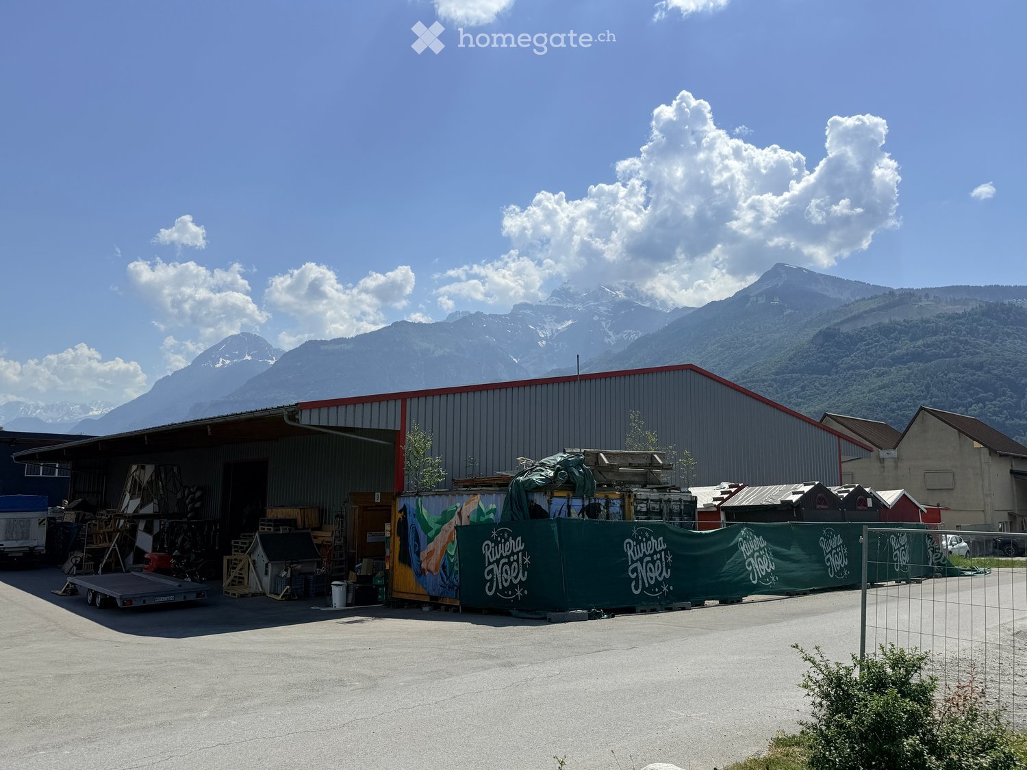 A large industrial-style building with a red roof and a green banner reading "Rivera Joie" in the foreground, surrounded by a parking area with various items and structures, with mountains in the background.