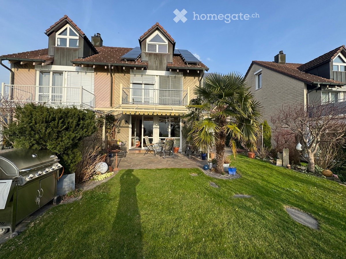 Well-maintained garden with a lawn, patio seating area, barbecue, and surrounding houses with balconies and solar panels.