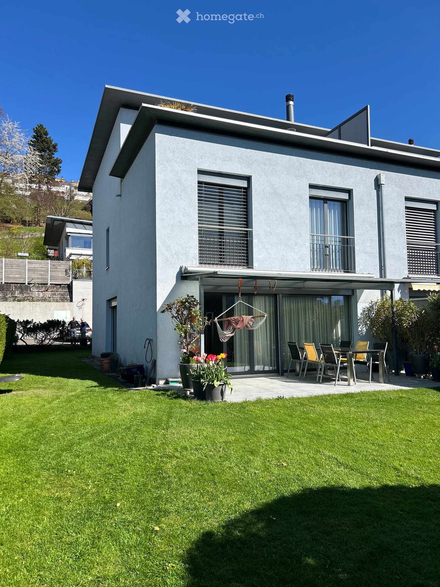 Modern two-story house with light gray exterior, large windows, and a covered patio area with outdoor furniture and potted plants.