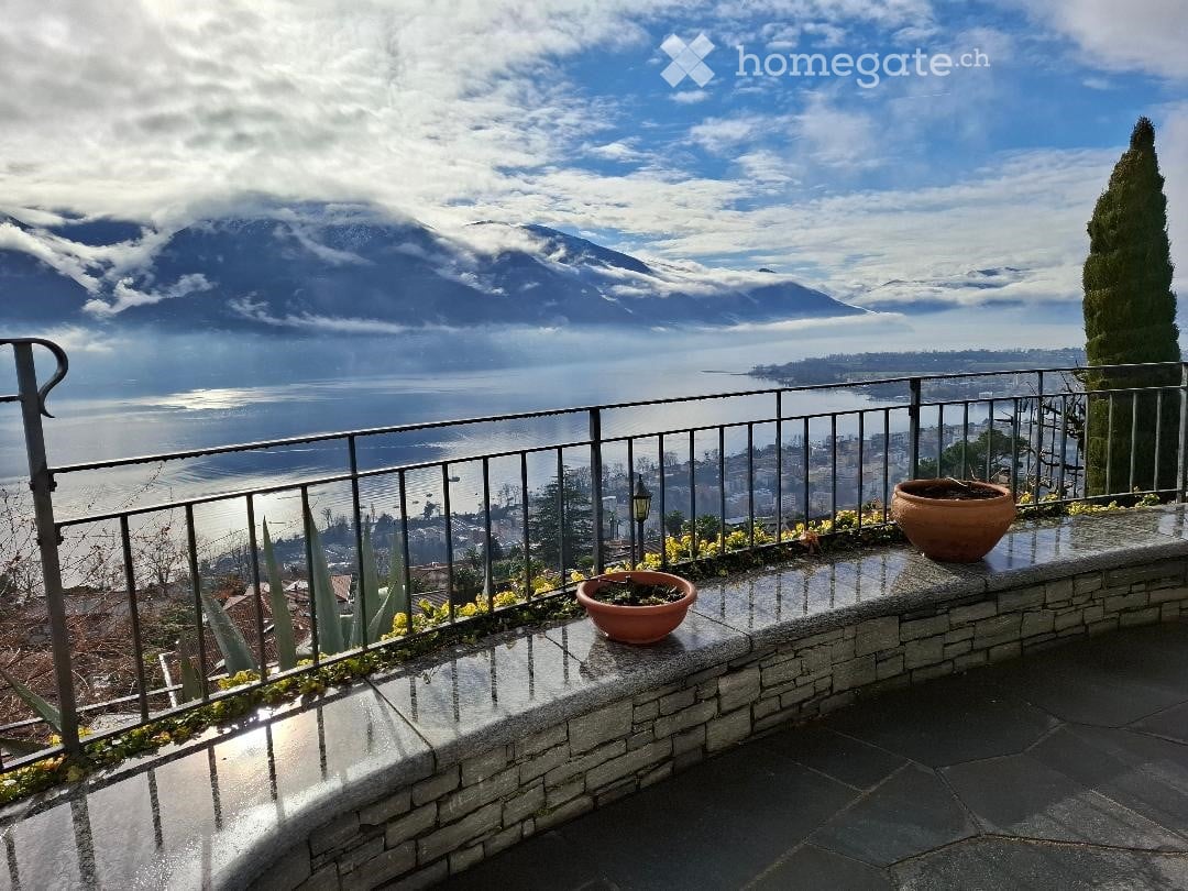 Terrace with black railings, concrete walkway, and potted plants, overlooking a sea and mountains