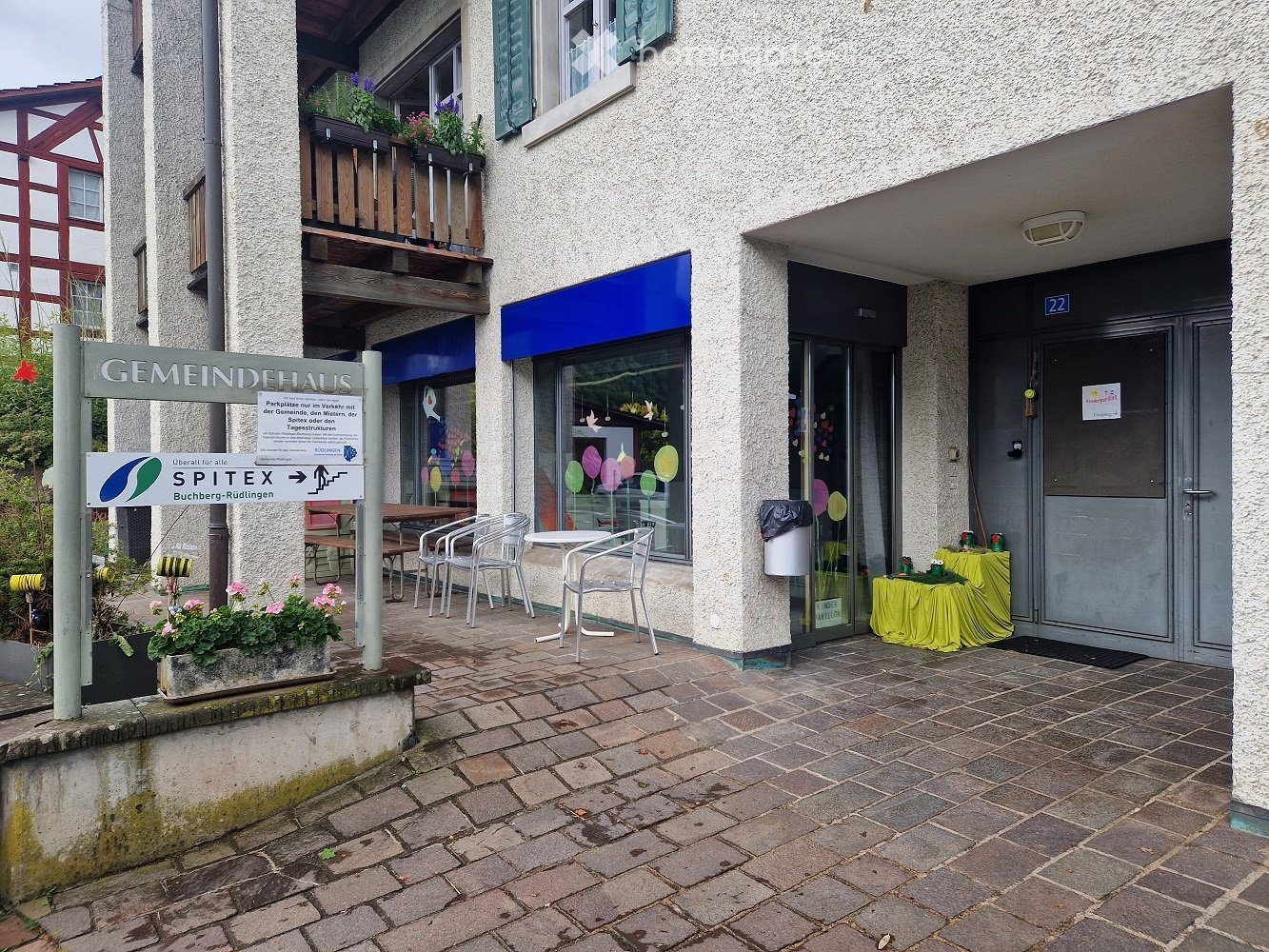 A small building with a sign reading "GEMEINDEHAUS" and a blue awning. There is a small outdoor seating area with tables and chairs, and a yellow tablecloth on a table near the entrance.
