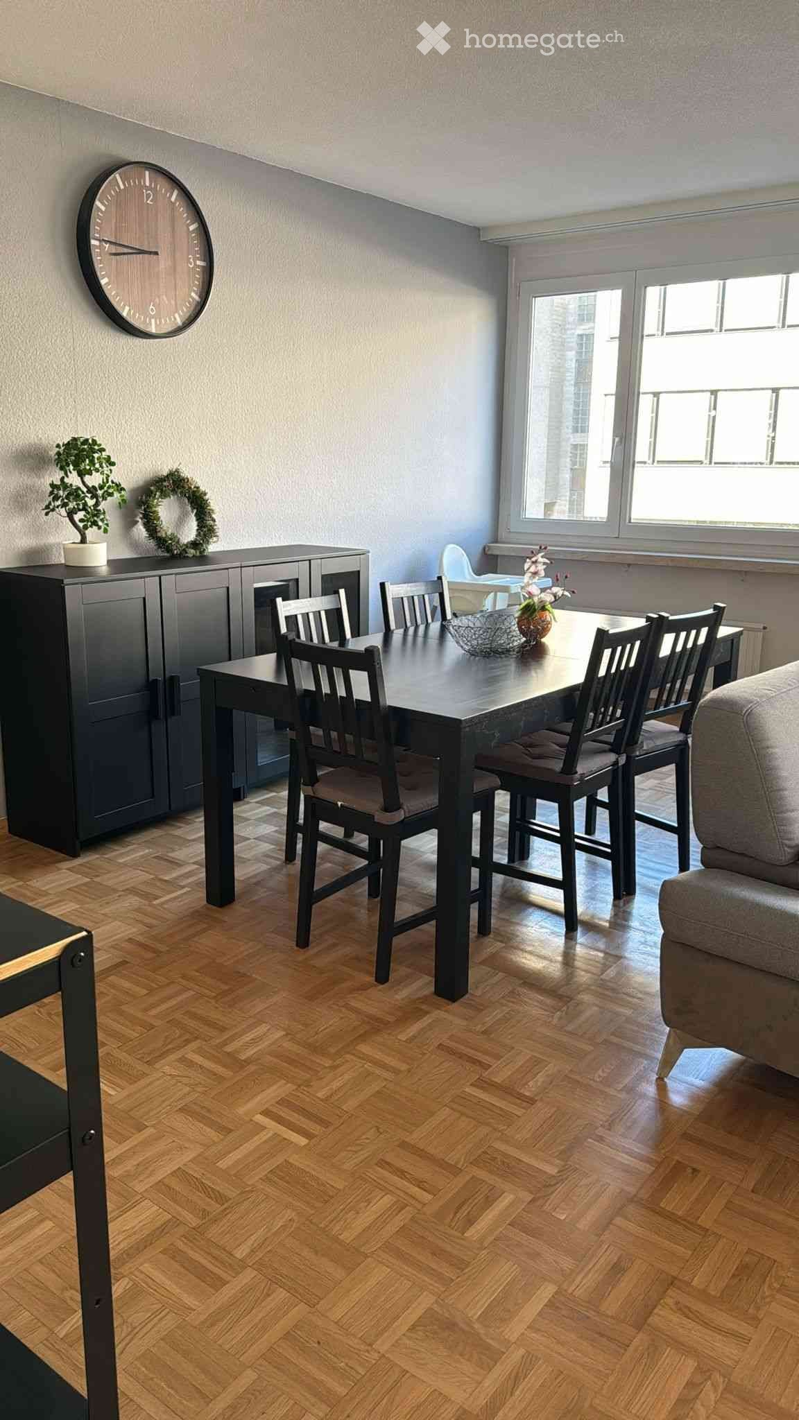 Dining area with a dark wooden dining table and chairs, a grey cabinet with a clock, a small plant, and a wreath on top, and a grey sofa visible in the corner.