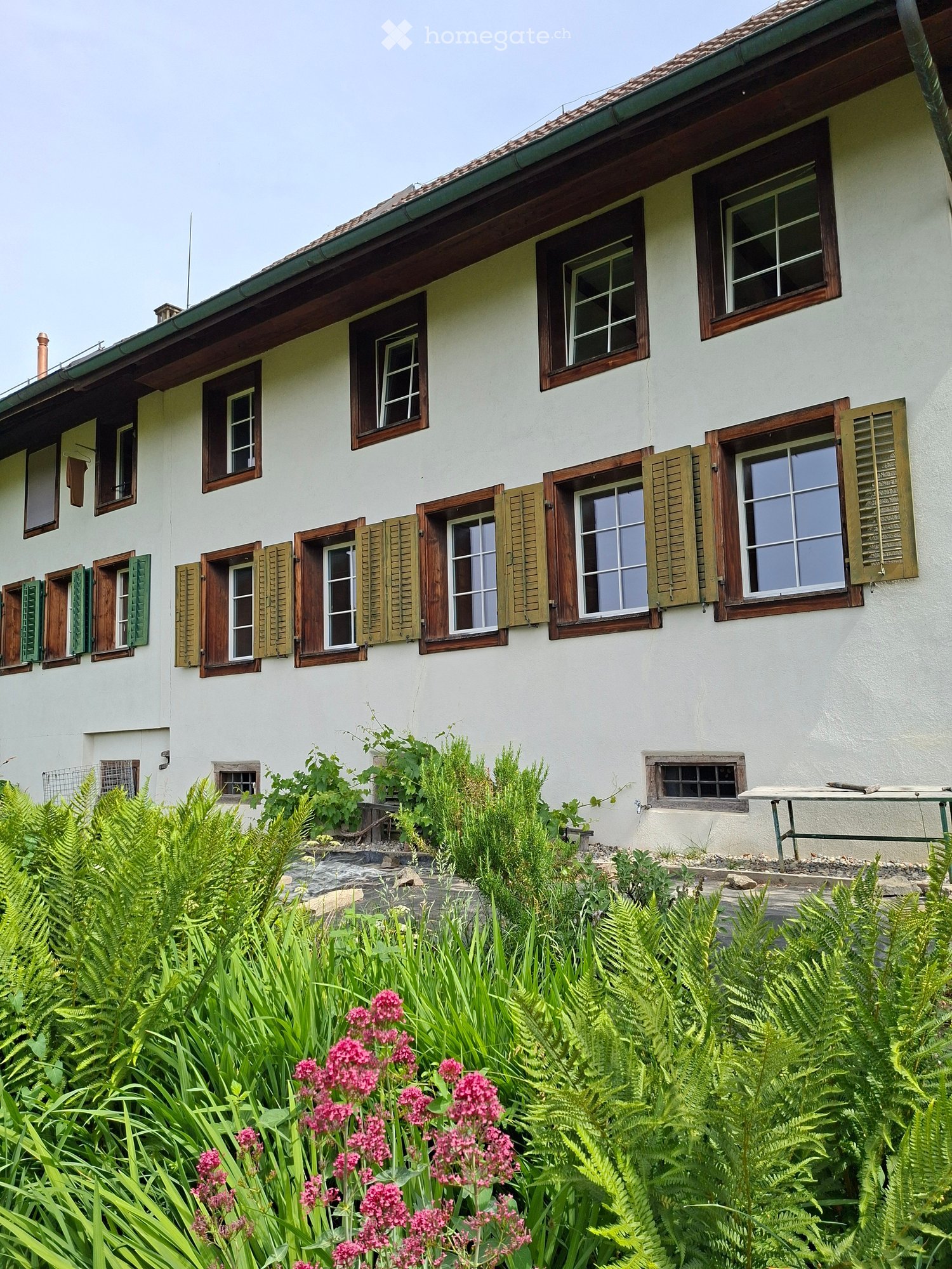 Two-story house with white walls, wooden shutters, multiple windows, and a well-maintained garden with ferns and pink flowers.