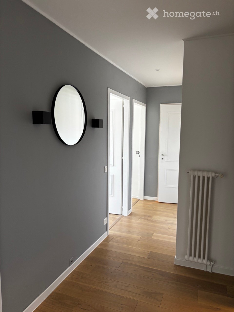 A modern hallway with grey walls, wooden flooring, and white doors leading to other rooms. Features a round wall mirror and a radiator on the right side.