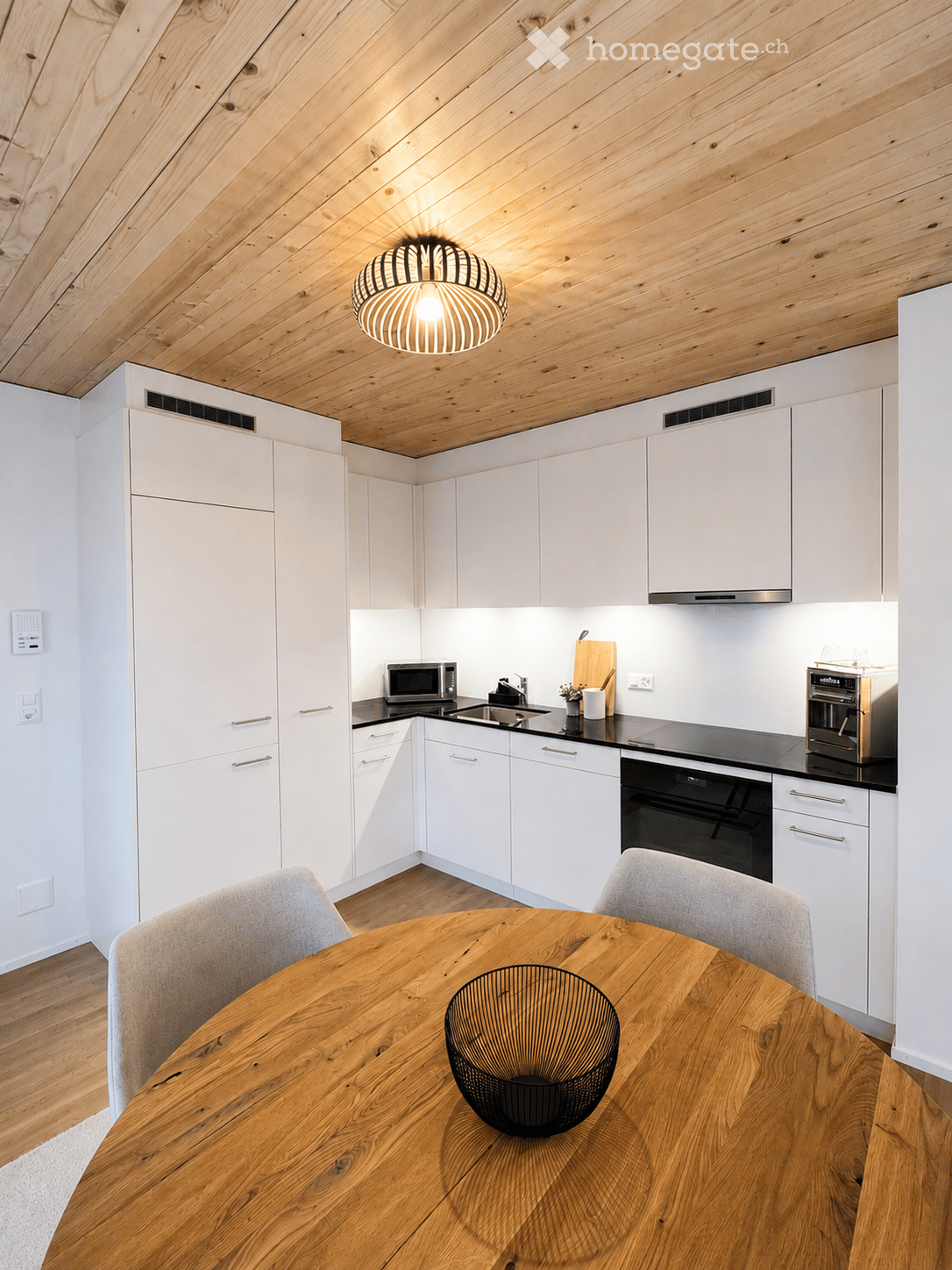 Modern kitchen with white cabinets, wooden table, and integrated appliances including microwave and oven. Features a sleek design with a pendant light and wooden flooring.