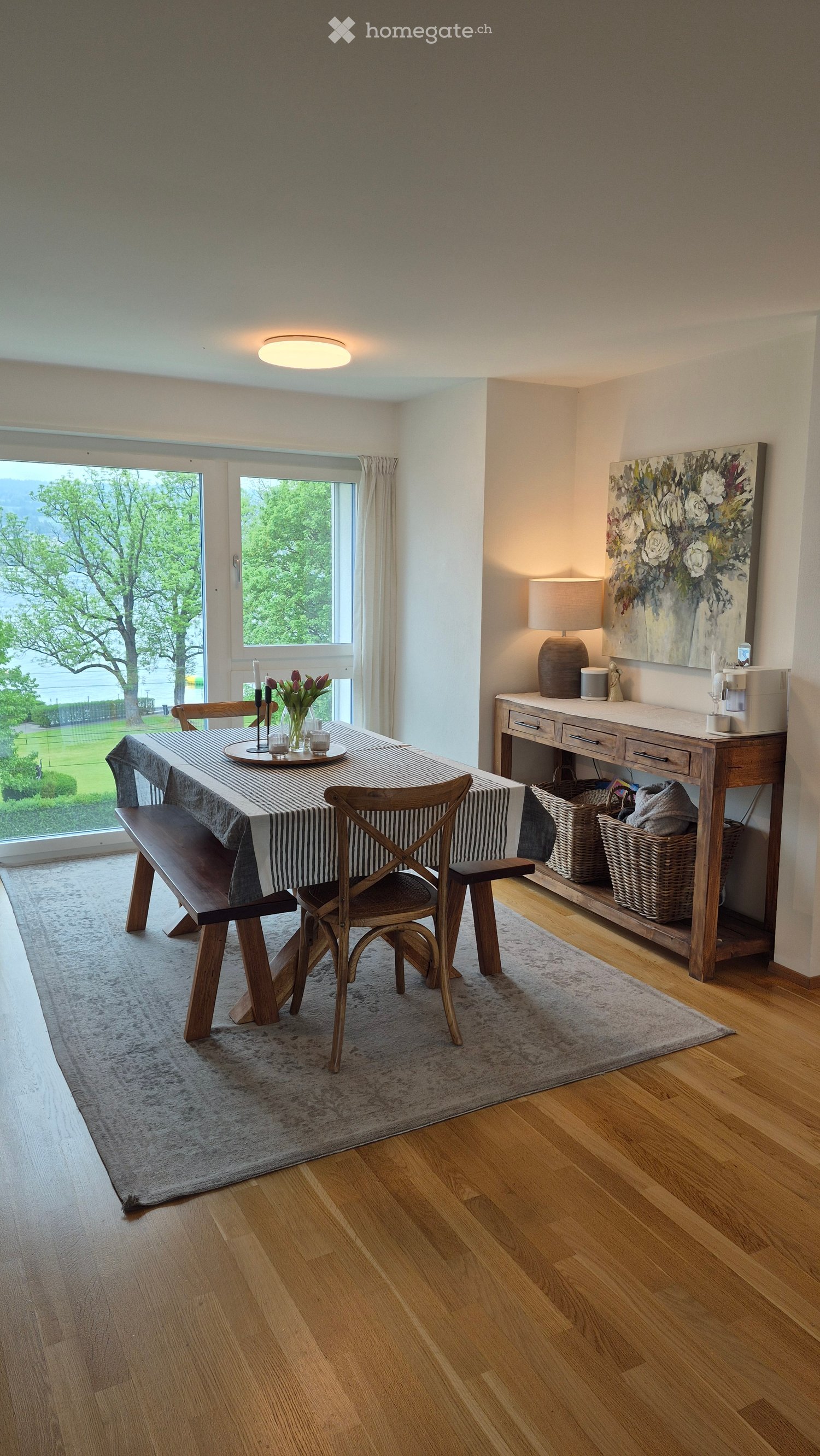 Bright dining area with wooden floor, large windows, a long wooden bench, a wooden chair, a sideboard with a lamp, a painting, and a vase with flowers on the table.