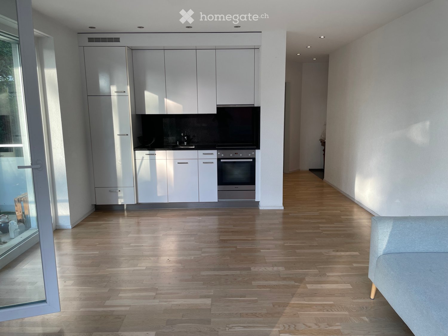 Modern, minimalist kitchen with white cabinets, stainless steel oven, and a sleek wooden floor. A grey sofa is visible in the adjacent living space.