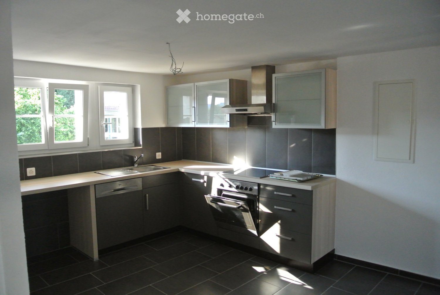 Modern kitchen with dark grey tiles, white countertops, stainless steel appliances, and large windows letting in natural light.