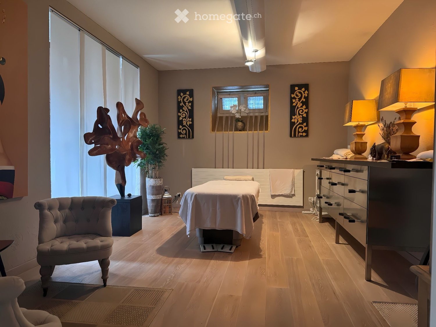 Well-lit, modern room with wooden flooring, a massage table, a plush chair, a side table with lamps, a decorative sculpture, and a potted plant.