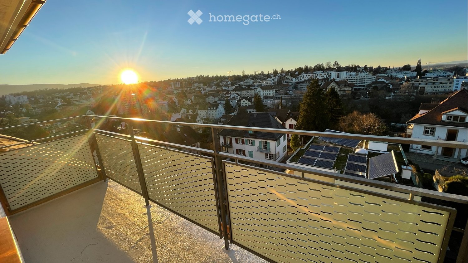 Balcony with metal railing and glass panels, overlooking a cityscape with houses and solar panels.