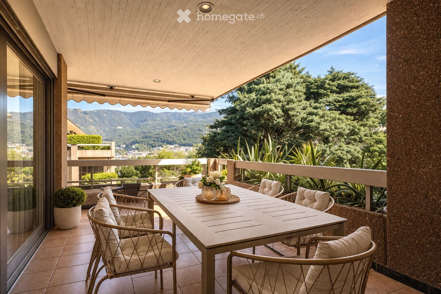 Balcony with a wooden table, four chairs, and a flower pot, overlooking a scenic view of trees and mountains.