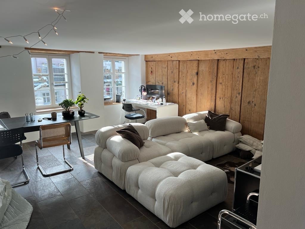 Modern living room with a large sectional sofa, coffee table, and a desk area with a screen. The room features wooden paneling on one wall and large windows letting in natural light.