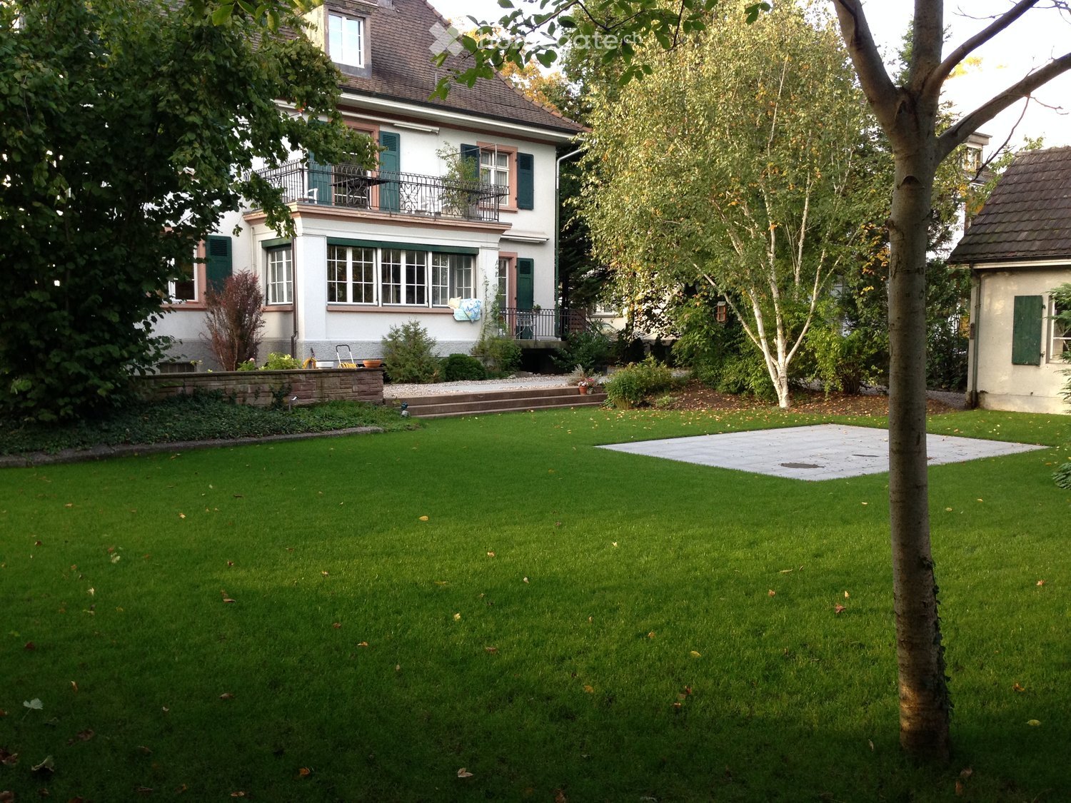 Well-maintained lawn with a tree, a small patio area, and a two-story house with a balcony and green shutters in the background.