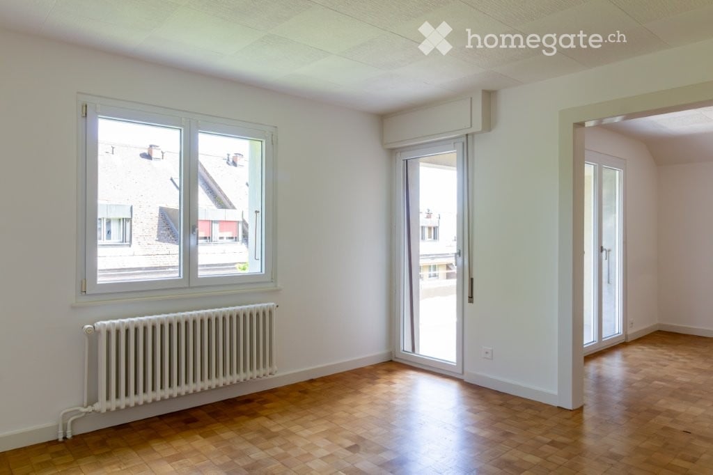 Empty room with wooden flooring, white walls, large windows, radiator, and a sliding glass door leading to another space.