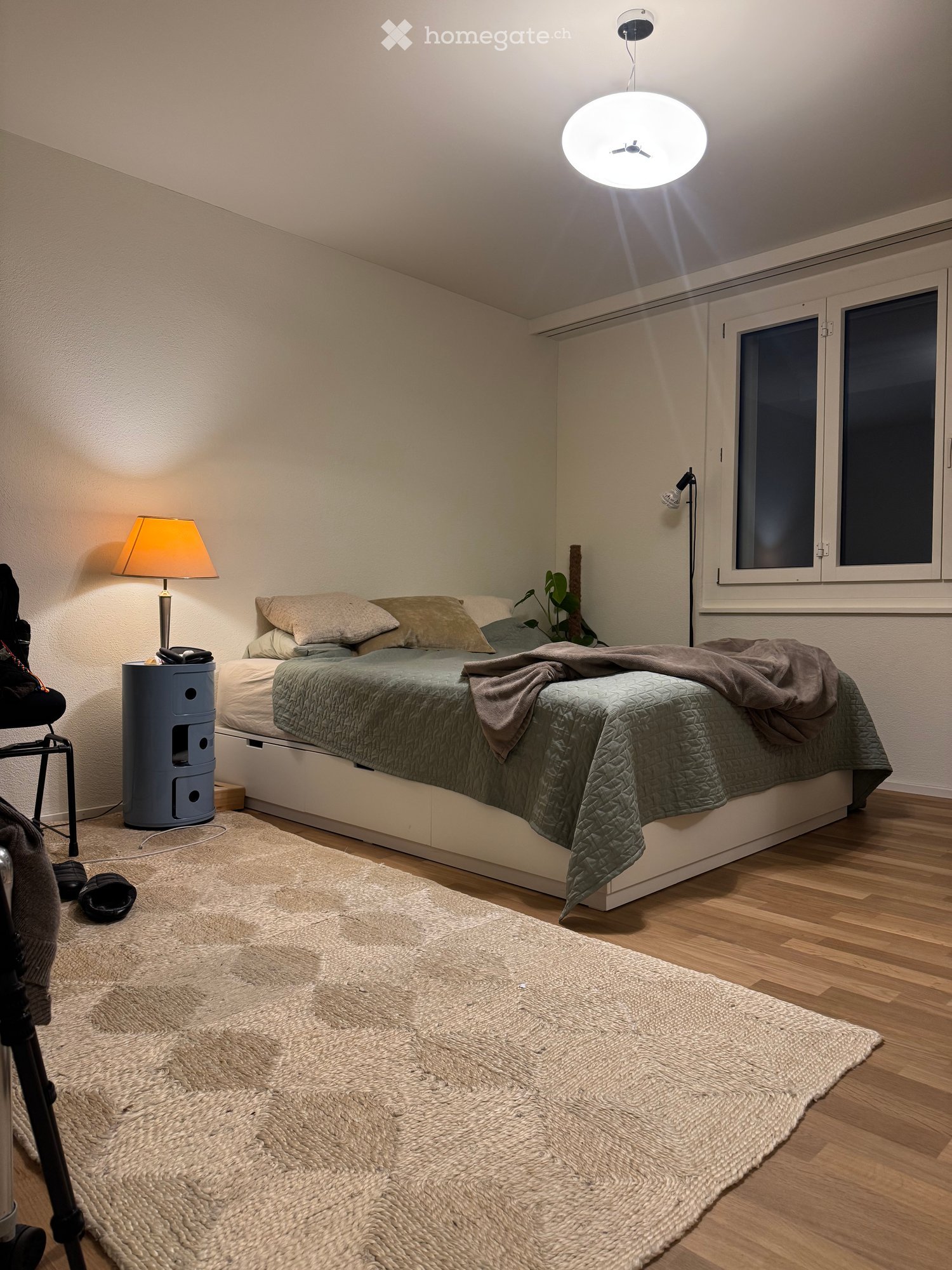 Cozy bedroom with a double bed, beige bedding, a bedside table with a lamp, a large round ceiling light, a woven rug, and a small plant near the window.