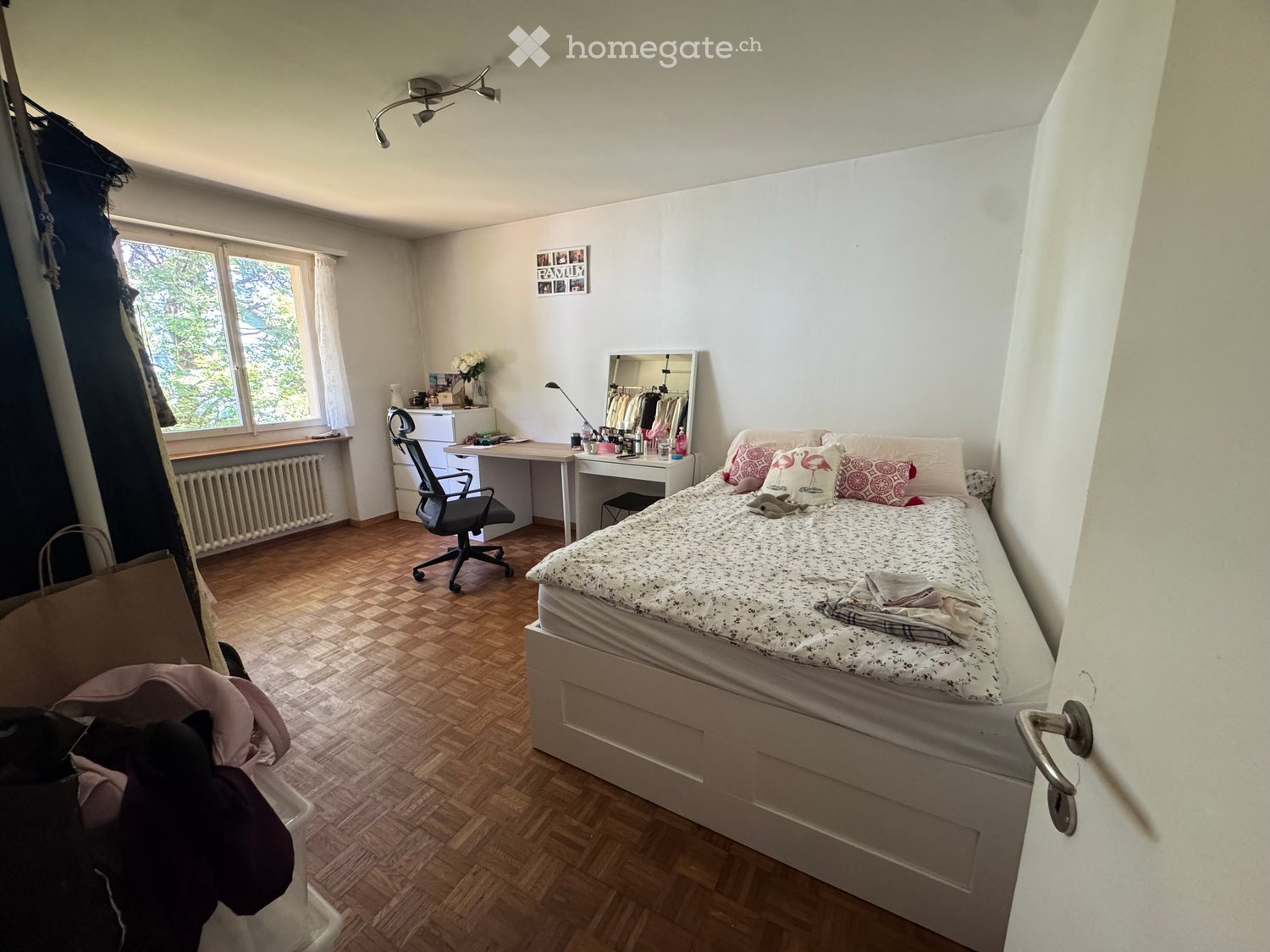 Cozy bedroom with wooden flooring, a single bed with pillows and a blanket, a desk with a chair, a mirror, and a radiator under the window.