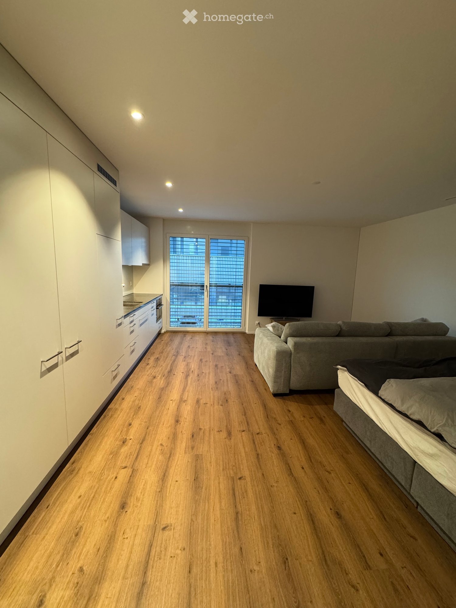 Open-plan living and sleeping area with wooden flooring, a grey sofa, a television, a bed, and modern kitchen cabinets.