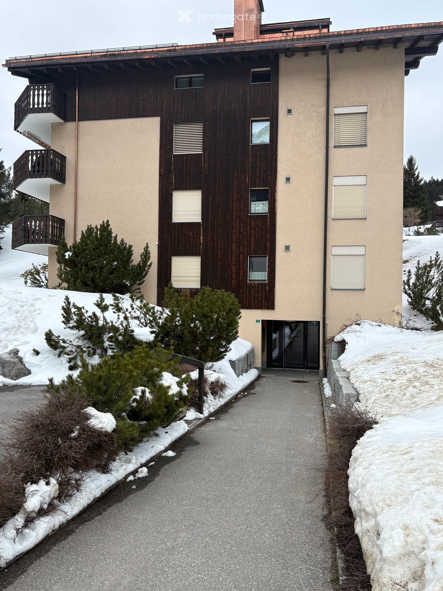 Two-story building with a wooden facade and beige walls, featuring multiple windows and balconies, surrounded by snow-covered ground and small evergreen trees.