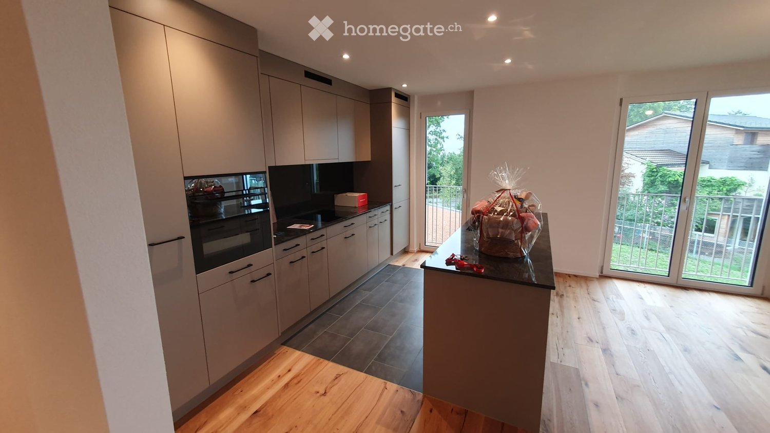 Modern kitchen with light grey cabinets, dark countertops, and a central island. Features an oven, a gift basket on the island, and large sliding glass doors leading to a balcony.