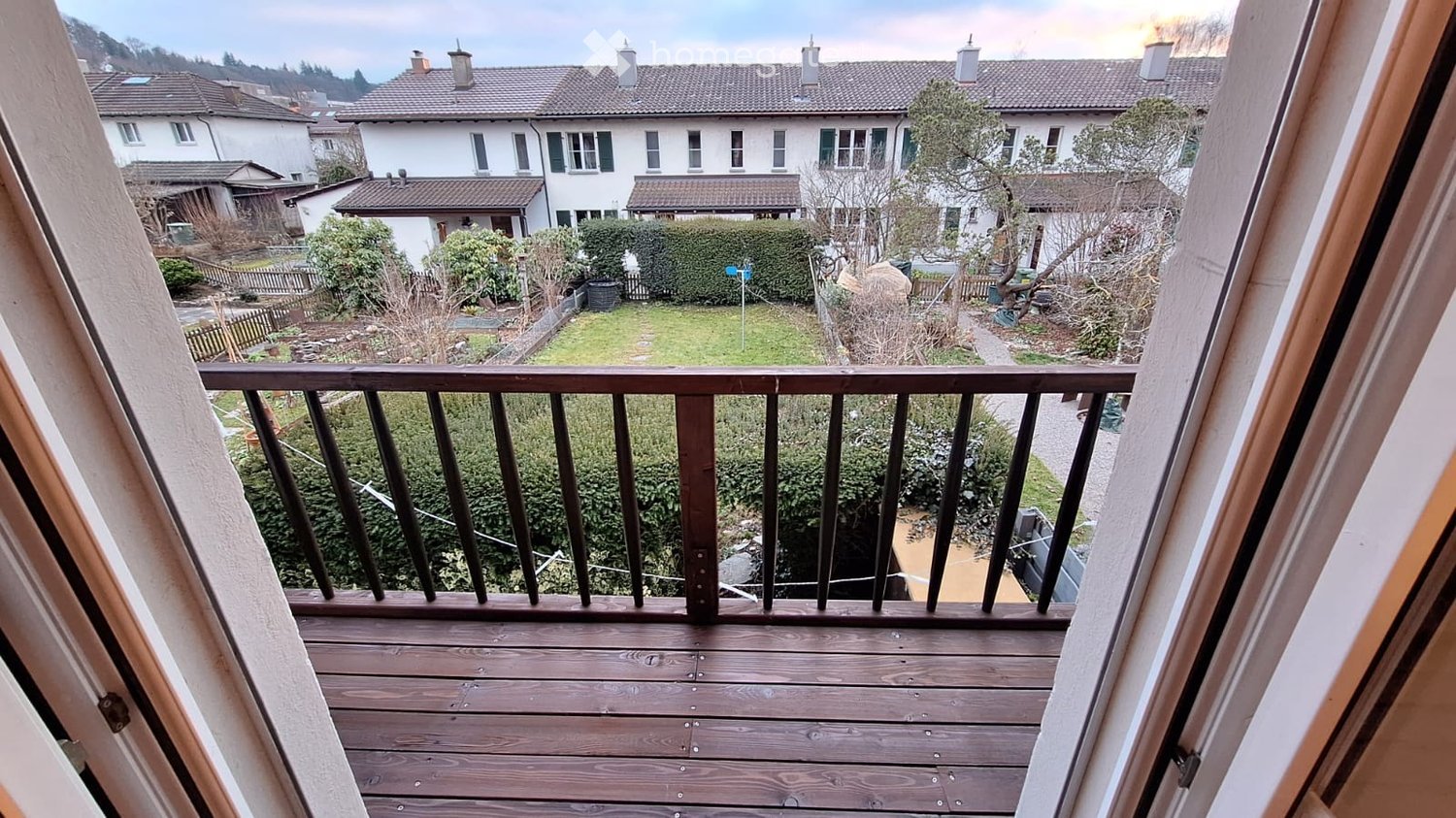 Wooden balcony with metal railing, overlooking a residential area with houses, greenery, and a pathway.