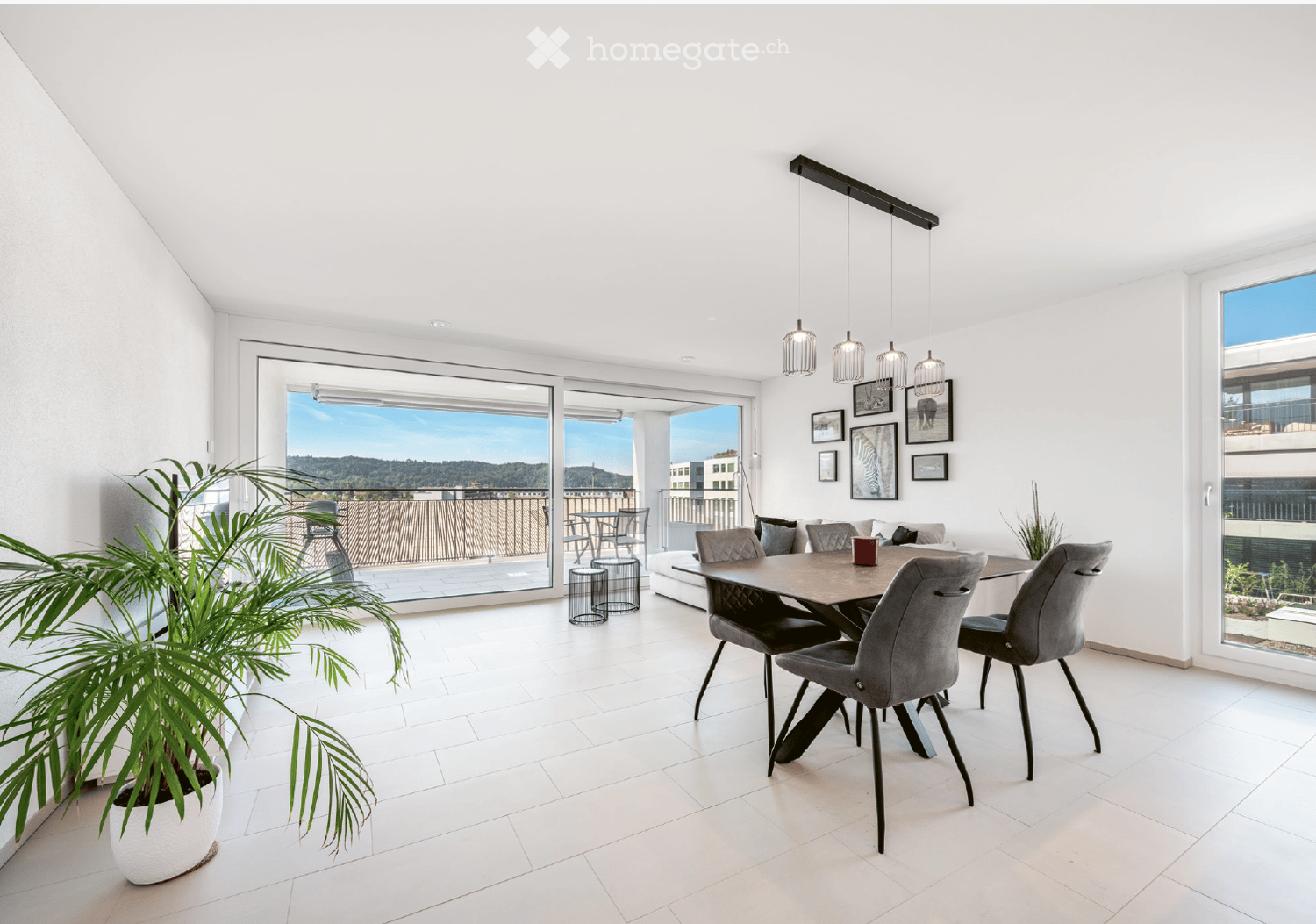 Modern dining area with white tiled flooring, large windows offering a scenic view, a dining table with four chairs, a sofa, and decorative picture frames on the wall.
