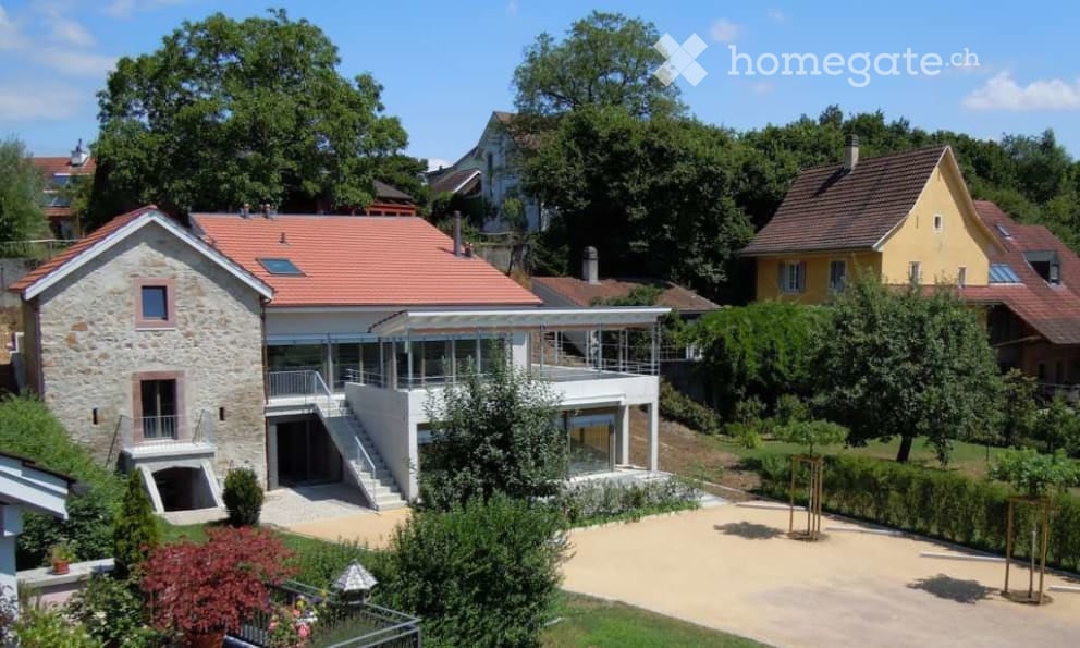 Modern two-story house with a stone facade and red-tiled roof, featuring a large glass-enclosed balcony area and a landscaped garden with trees and shrubs.