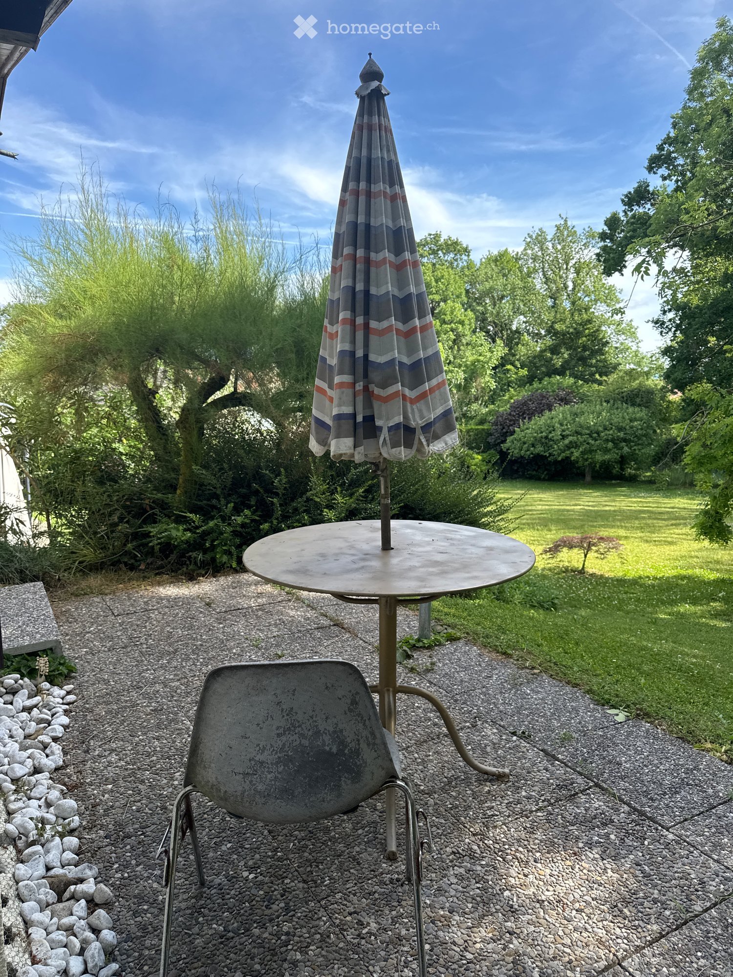 Garden area with a round metal table, a single chair, and a closed umbrella, surrounded by gravel and greenery.