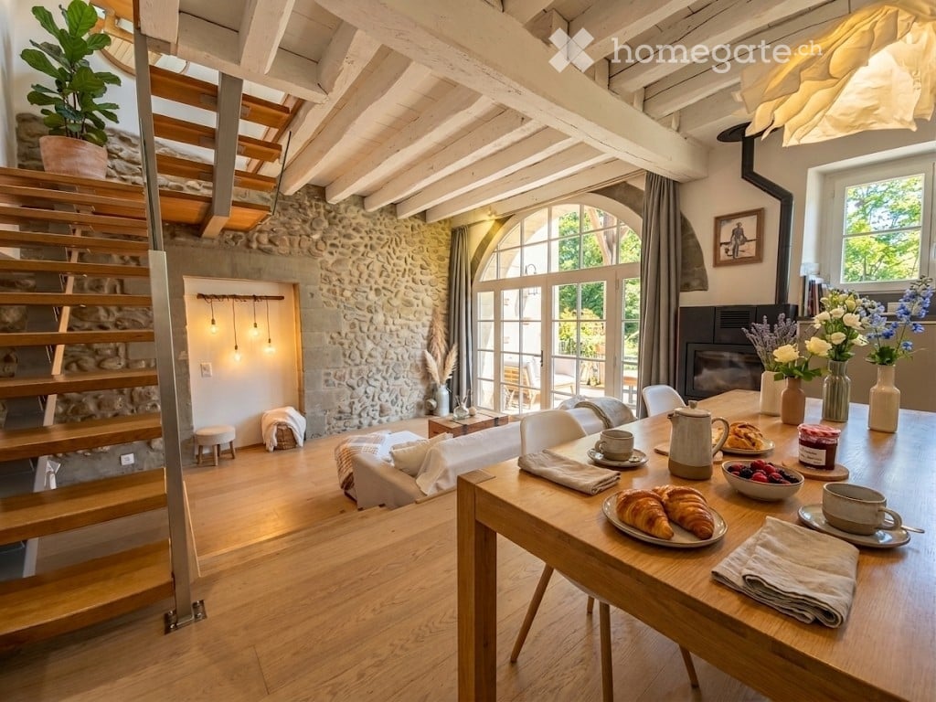 Bright, open-plan living and dining area with exposed wooden beams, stone walls, large windows, and a fireplace. Features a sofa, coffee table, dining table with chairs, and decorative vases.