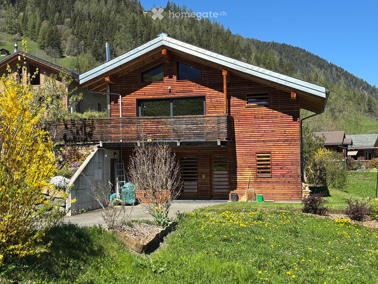 A two-story wooden house with a modern design, featuring large windows, a balcony, and a well-maintained garden with colorful flowers and shrubs.