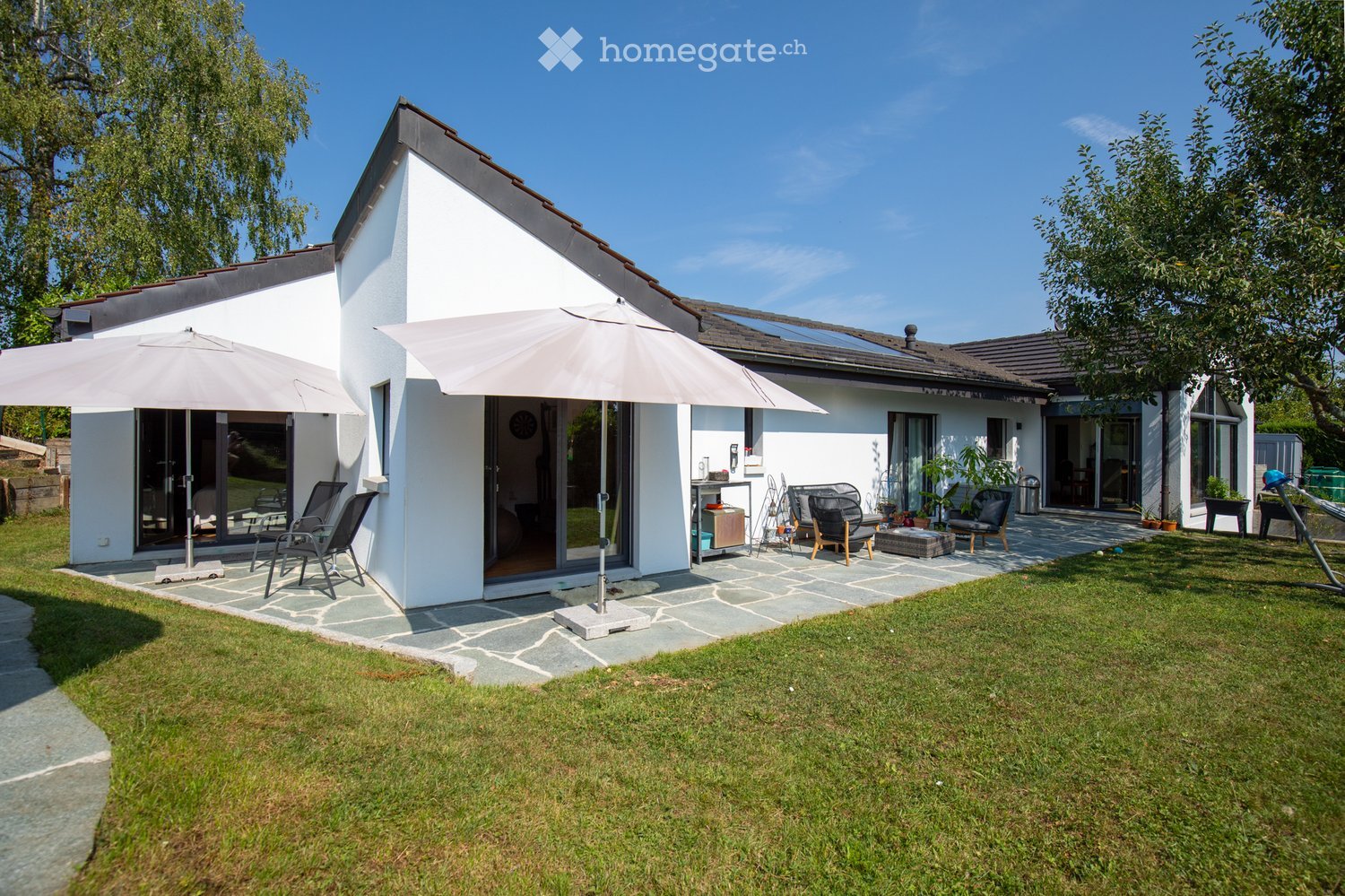A modern white house with a well-maintained garden, featuring outdoor seating, umbrellas, and a stone patio.