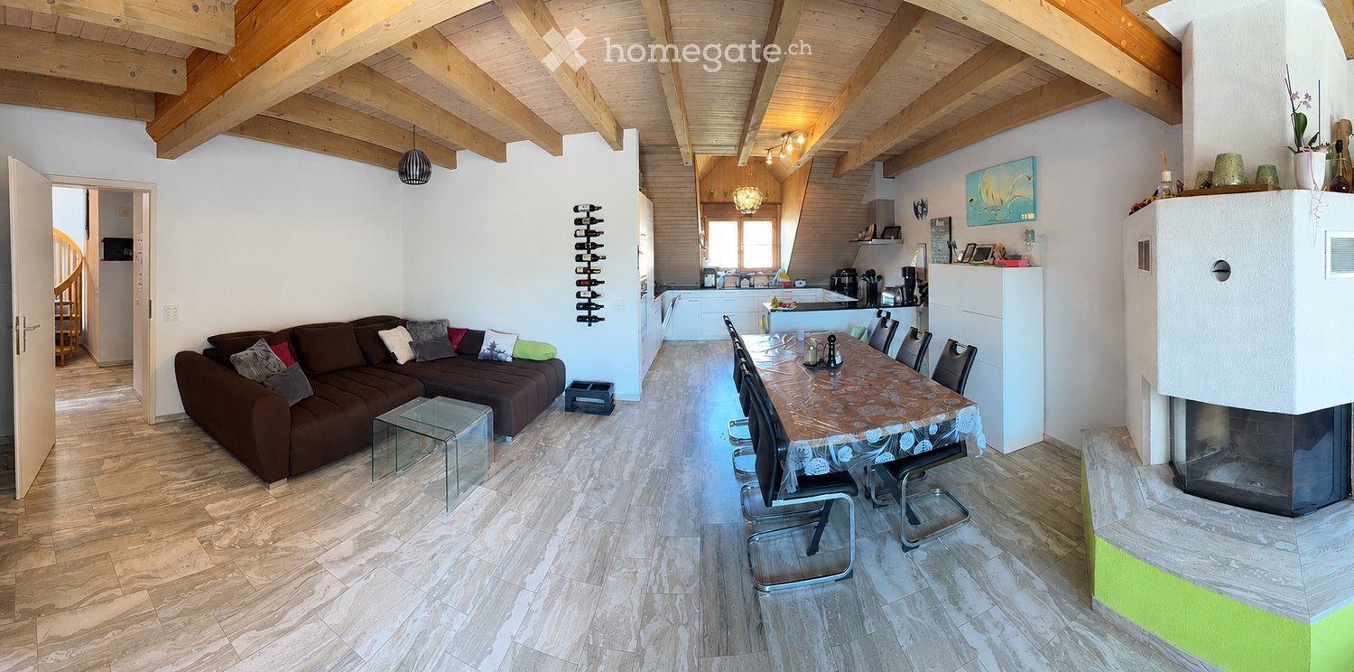 Open-plan living and dining area with exposed wooden beams, a brown sectional sofa, a glass coffee table, a dining table with chairs, and a modern fireplace. The kitchen area features white cabinets and stainless steel appliances.