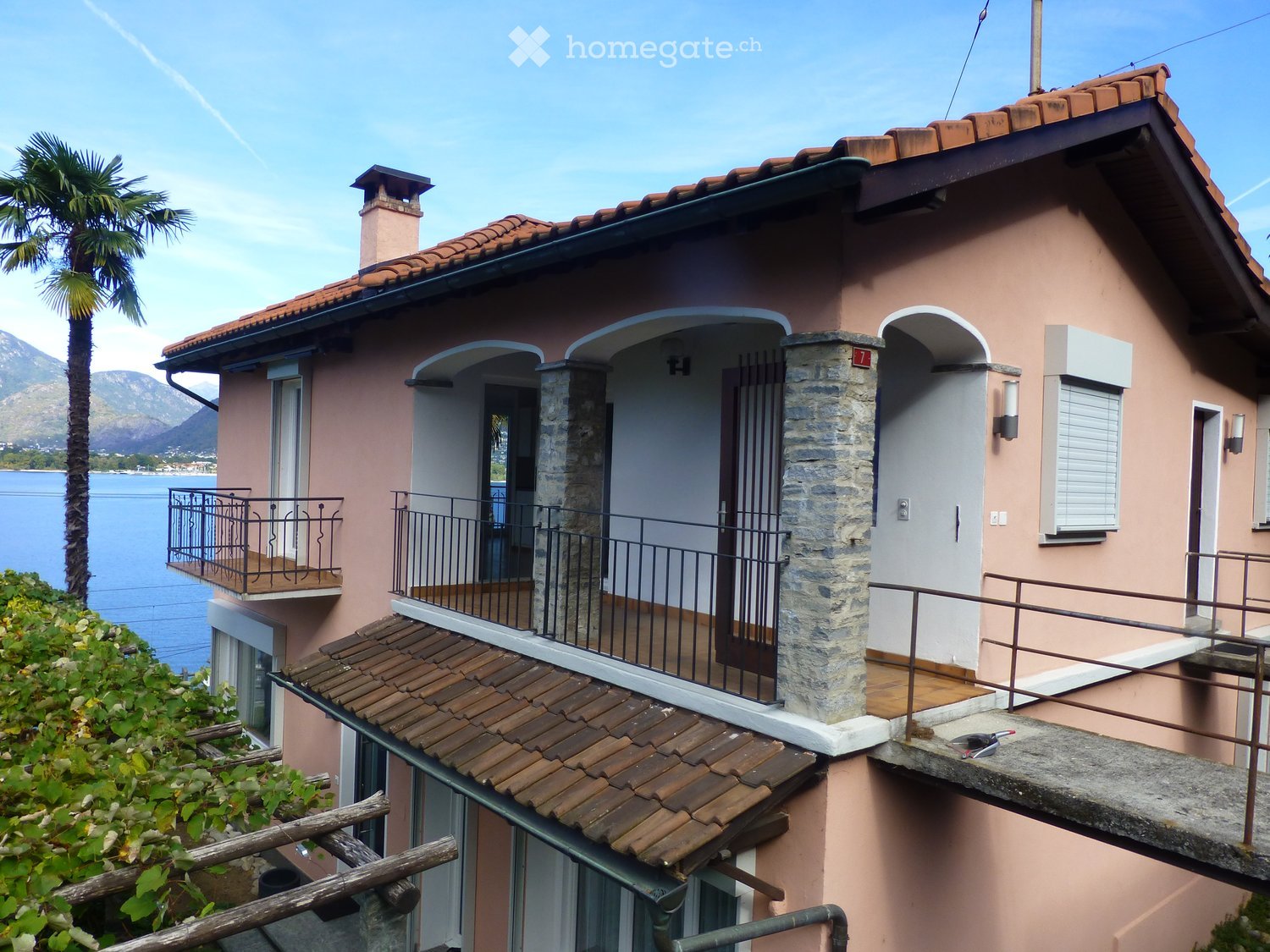 Two-story house with pink walls, terracotta roof, balconies, and a view of the lake and mountains.